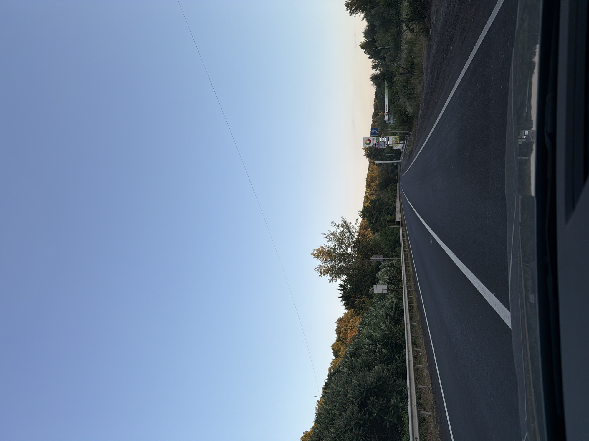 The image shows a road in the city of Velyki Lazy, Ukraine. The road is asphalt and has white lines marking the lanes. There are trees and bushes on either side of the road. In the distance, there is a gas station with a sign that says "amic." The sky is clear and blue. The sun is setting, casting a warm glow over the scene.