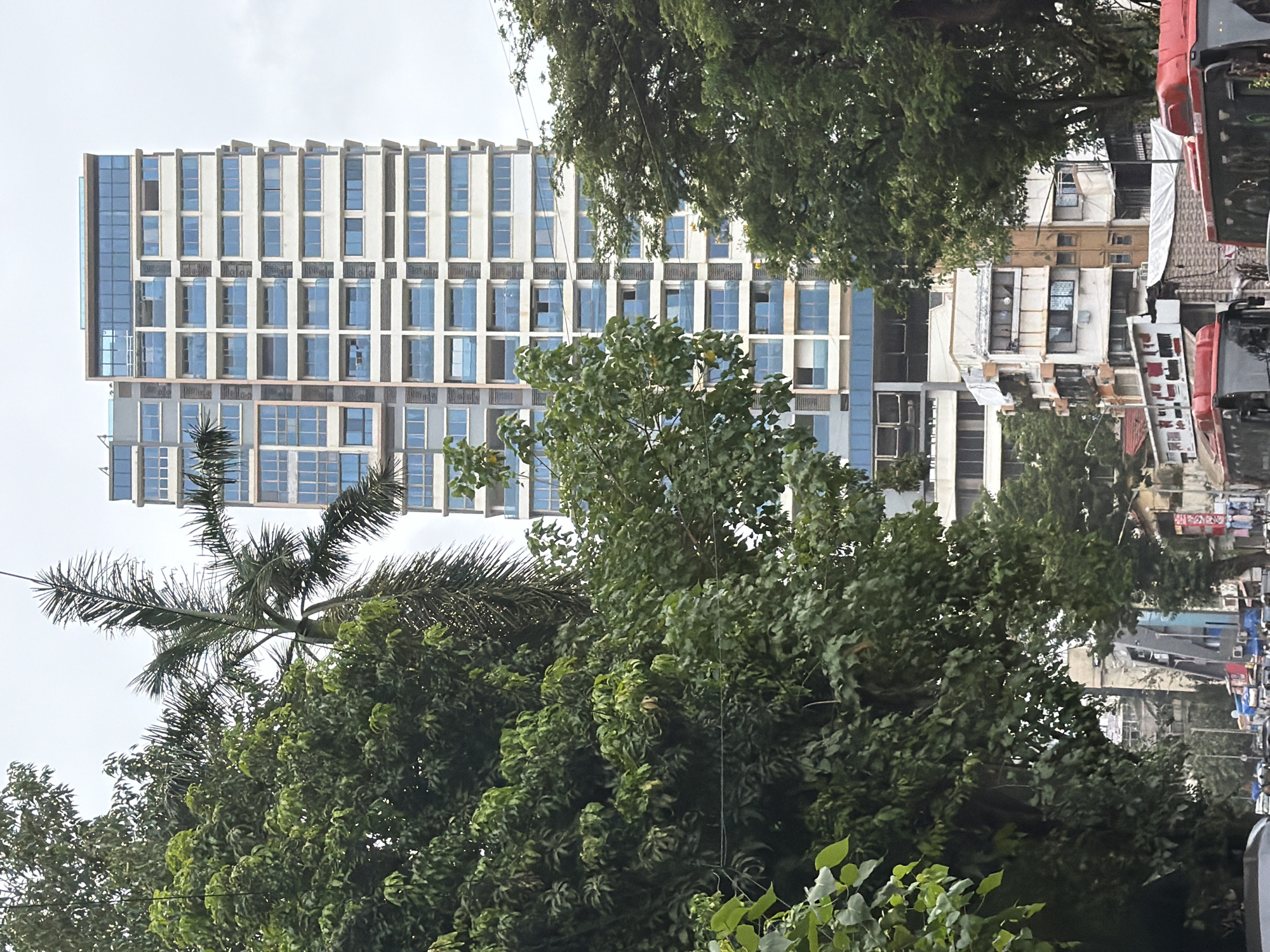 This is a medium-shot, eye-level, vertical, outdoor, daytime photograph of a street scene in Mumbai, India, with a focus on a tall, modern, white and blue-windowed residential building. Lush green trees, including a tall palm tree, dominate the foreground and frame the right side of the image, partially obscuring the view of the residential building. The building features a grid-like facade of windows and balconies. Below the tall building, older, lower-rise buildings with a mix of architectural styles are visible, some appearing to be commercial establishments with signage. A street with multiple lanes is apparent, with several red buses visible in the foreground, suggesting a busy urban environment. The sky is overcast, indicating a cloudy day. The image captures the contrast between modern high-rise architecture and older urban structures, characteristic of many parts of Mumbai.