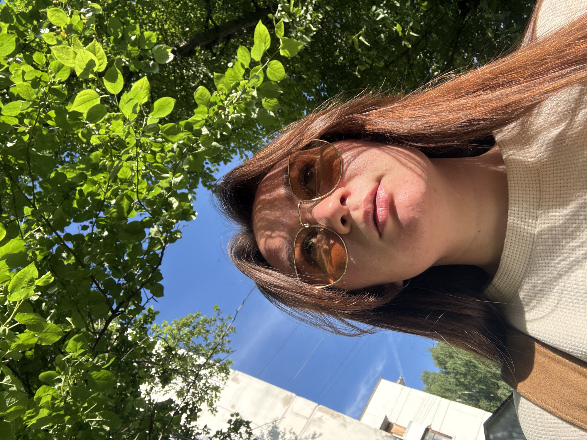 This low-angle outdoor photograph features a person, likely female, captured from an upward perspective against a backdrop of trees and sky. The individual, positioned in the lower center-right of the frame, has long, rich reddish-brown hair. She wears amber-tinted, round sunglasses with thin gold frames, which obscure most of her eyes. Her lips are slightly parted, and her expression is neutral. Strong, dappled shadows from the overhead tree canopy fall across her face and hair, indicating direct sunlight. She is wearing a light-colored, possibly beige, waffle-knit top, and a segment of a brown garment or strap is visible near her shoulder.
The upper and left portions of the image are dominated by the vibrant green foliage of trees, with sunlight illuminating the leaves and creating distinct shadows. A clear, bright blue sky is visible between the leaves and behind the person, with a few faint cirrus clouds in the distance. In the lower background, parts of light-colored buildings with windows are visible, suggesting an urban or suburban setting. The bright illumination indicates it is daytime, likely mid-morning or afternoon, on a sunny day. The scene depicts an outdoor moment in Chaikovsky, Russia. No visible text is present.