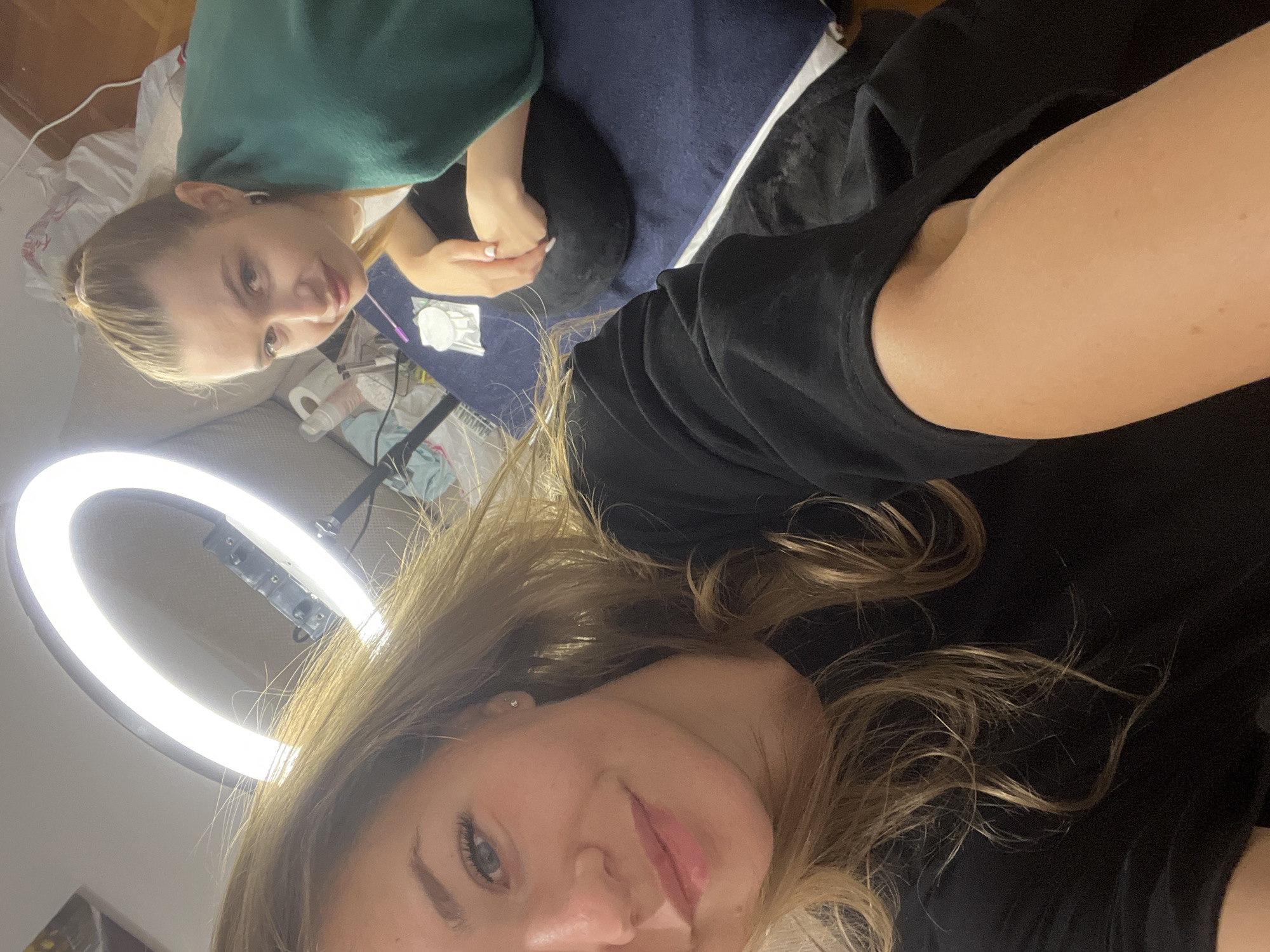 Two Caucasian females are in a room. One is in the foreground, wearing a black t-shirt, with shoulder-length blonde hair. Her head is oriented toward the lower part of the image. Above her is a ring light. The second female is partially visible in the upper left corner of the frame, wearing a green sweater. She is sitting at a table with a dark blue surface, where a disposable medical needle is lying. The image appears to be a selfie. The location is Budva, Montenegro.