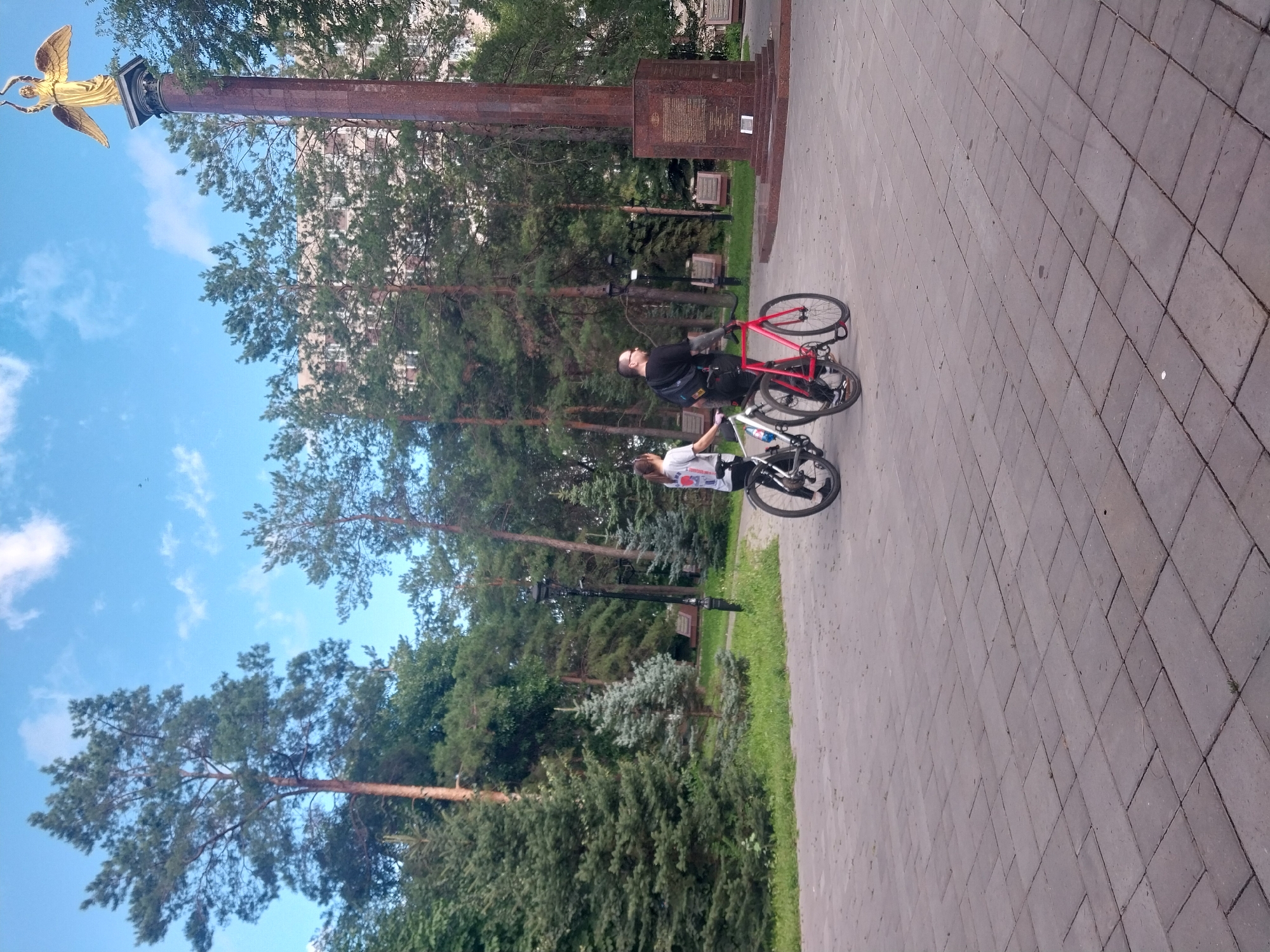 Two people, a man and a woman, are riding bicycles on a paved walkway in a park. The man is wearing a black t-shirt and glasses, and the woman is wearing a white t-shirt with graphics. They are riding side-by-side, with the man on a red bicycle and the woman on a white bicycle. In the background, there is a tall, ornate monument with a golden angel statue on top, surrounded by trees and a building. This monument is located in the Victory Park (Park Pobedy) in Orenburg, Russia. The sky is blue with scattered clouds. The walkway is made of grey rectangular paving stones.