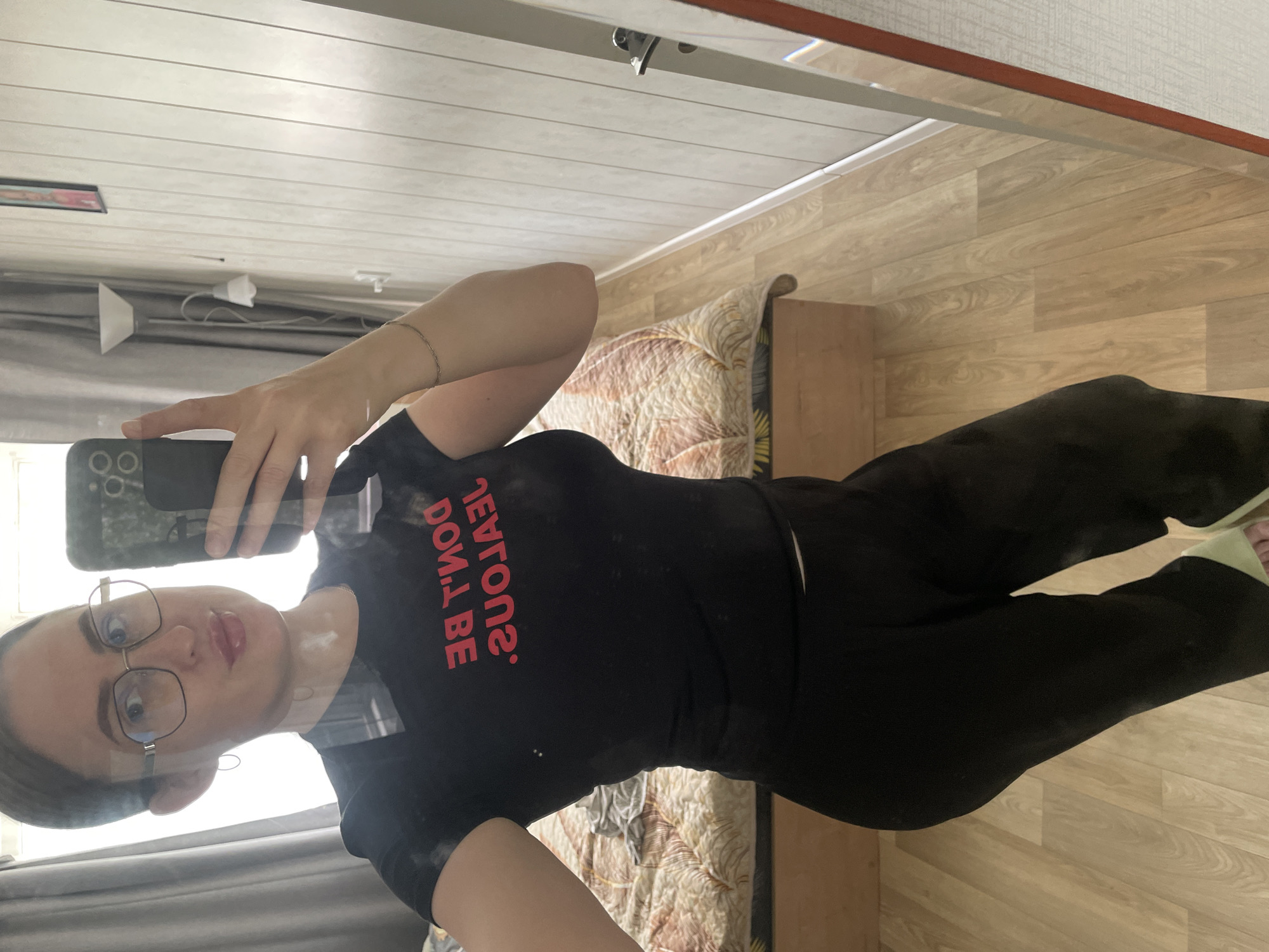 The image shows a full-body mirror selfie of a woman standing in what appears to be a bedroom. She is wearing a black t-shirt with red text that reads "DON'T BE JEALOUS." and black pants. She has on glasses and is holding a phone in her right hand. The background includes a bed with a patterned comforter, a mirror, a floor lamp, and wood-paneled walls. The flooring is light-colored wood laminate. The overall impression is that of a casual, indoor setting. There is no specific information in the image to suggest a location in the city of Сырково, Russia.