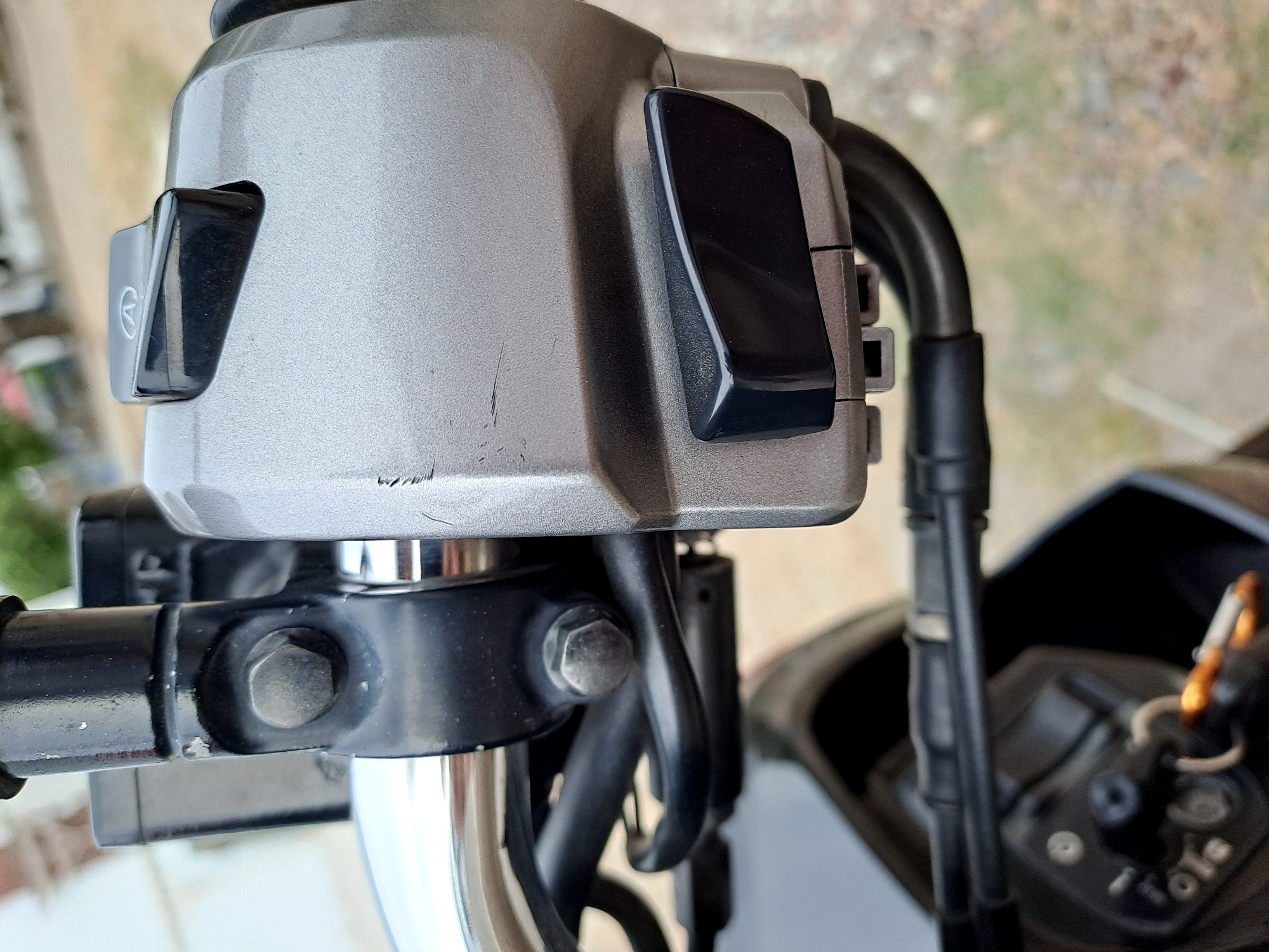 A close-up, eye-level, medium shot captures the right-hand handlebar assembly of a motorcycle. The handlebars are chrome, and attached to them is a grey plastic housing that contains two black switches. The upper switch has a small circular symbol with a diagonal line through it. The lower switch is rectangular with rounded corners. The housing shows some light scuff marks. Below the handlebar assembly, a black plastic component of the motorcycle is visible, along with a dark-colored key ignition. The background is out of focus, showing a blurred mix of green and brown, suggesting an outdoor environment. There are no people or animals present in the image. The scene is static and shows no activity.