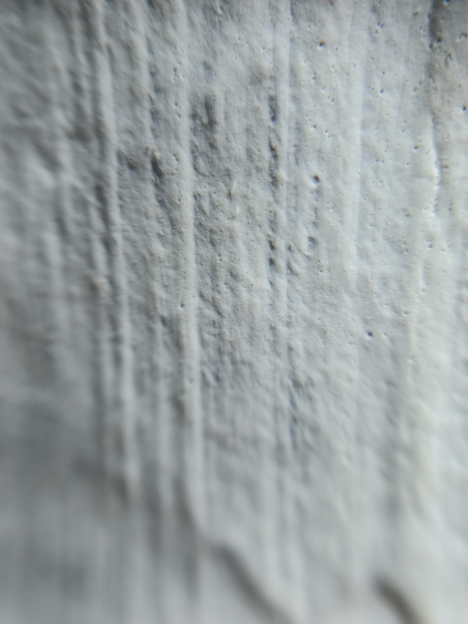 The image is a close-up, macro photograph of a textured, light-colored surface. The surface appears to be rough and uneven, with visible striations and small pockmarks or pores. The lighting creates subtle shadows that accentuate the texture. The overall impression is of a natural or man-made material with a weathered or unfinished quality. There are no people, recognizable objects, or environmental context provided that would place it in Margajaya, Indonesia, or any other specific location. The image focuses solely on the abstract qualities of the surface's texture.