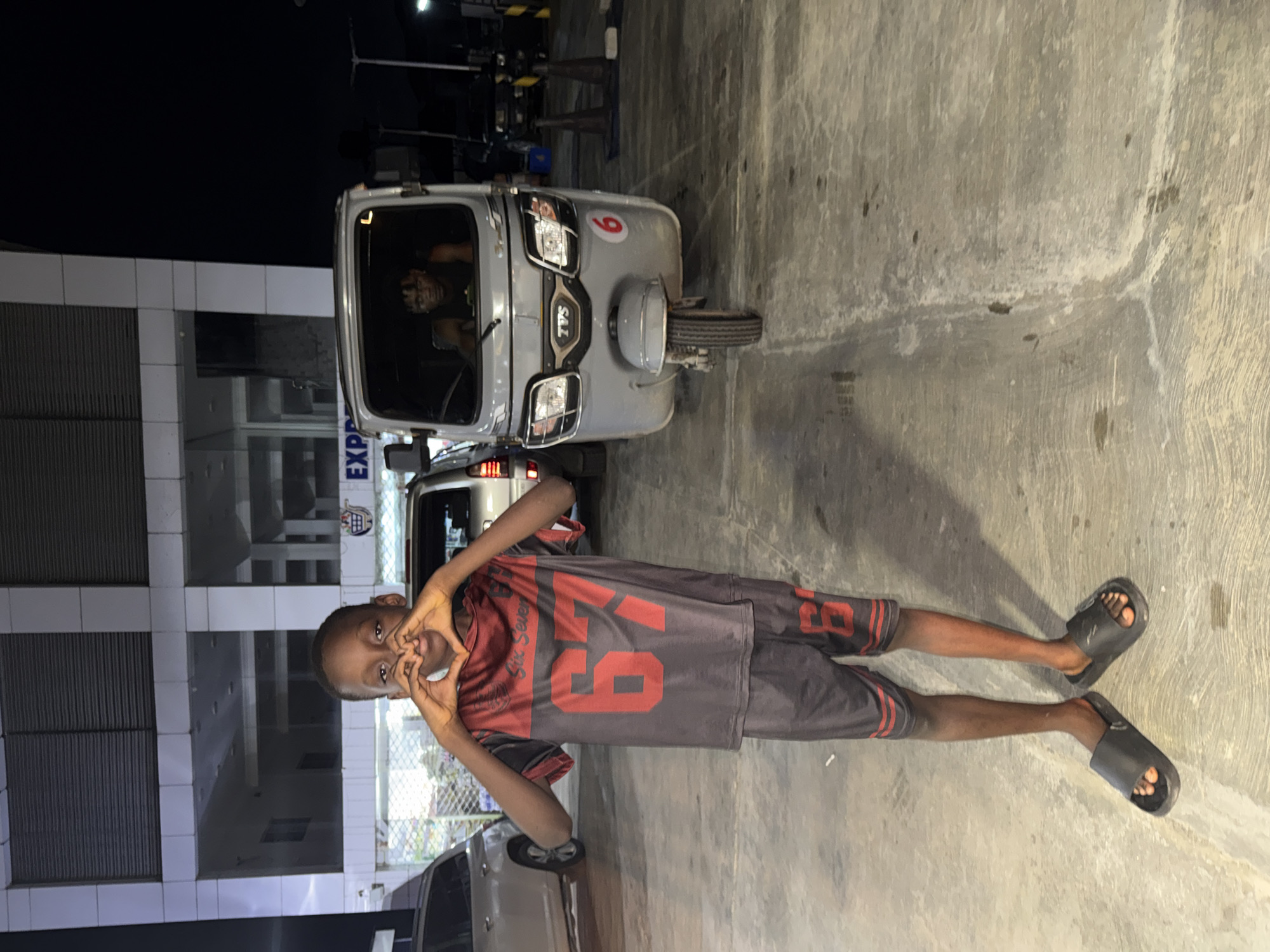 The image depicts an outdoor night scene in the city of Ado, Nigeria, featuring two individuals and several vehicles in front of a commercial building.
In the foreground, a young dark-skinned male, appearing to be a pre-teen, stands centrally, facing the camera. He is wearing a dark grey/black and reddish-orange athletic two-piece set, consisting of a short-sleeved shirt with "Six Seven" and a large "67" printed on the chest, and matching shorts with "67" on the left leg. He wears black slide sandals. His hands are positioned in front of his chest, forming a heart shape, and he is looking directly at the camera.
To the right of the boy, a light grey three-wheeled vehicle, specifically a TVS brand rickshaw (keke), is parked. A red circular sticker with the number "6" is affixed to its front right side. An adult dark-skinned male with short dark hair is visible through the windshield, seated in the driver's seat, looking towards the camera. He is wearing a dark sleeveless top.
In the background, a multi-story building with a white/light grey facade and numerous windows is visible. A sign partially reads "EXPR" with an associated logo, suggesting a commercial establishment. Artificial lighting illuminates the building's exterior and interior, contrasting with the dark night sky.
To the left of the boy, a silver-colored SUV or car is partially visible. The ground is a paved concrete surface, showing signs of wear and minor staining. Dark poles or bollards are present in the far right background, behind the three-wheeled vehicle.