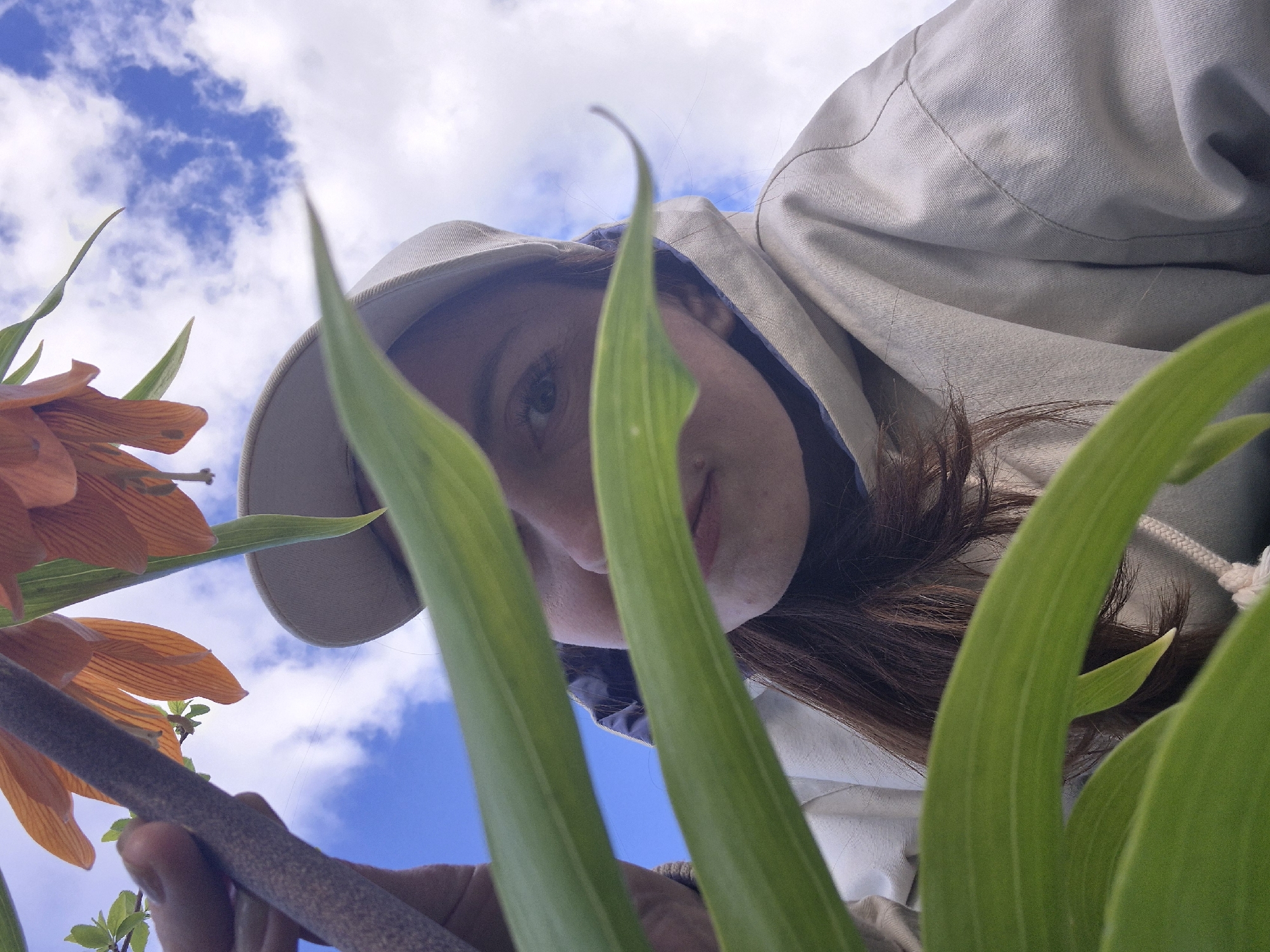 A low-angle shot depicts a person wearing a hooded jacket and a light-colored cap, looking downward towards the camera. The subject's face is partially obscured by long, green leaves in the foreground. An orange flower is visible on the left side of the frame, with a blue sky and white clouds in the background. A hand, holding the stem of the flower, is visible in the lower left corner. The location is presumed to be in the city of Chaikovsky, Russia.