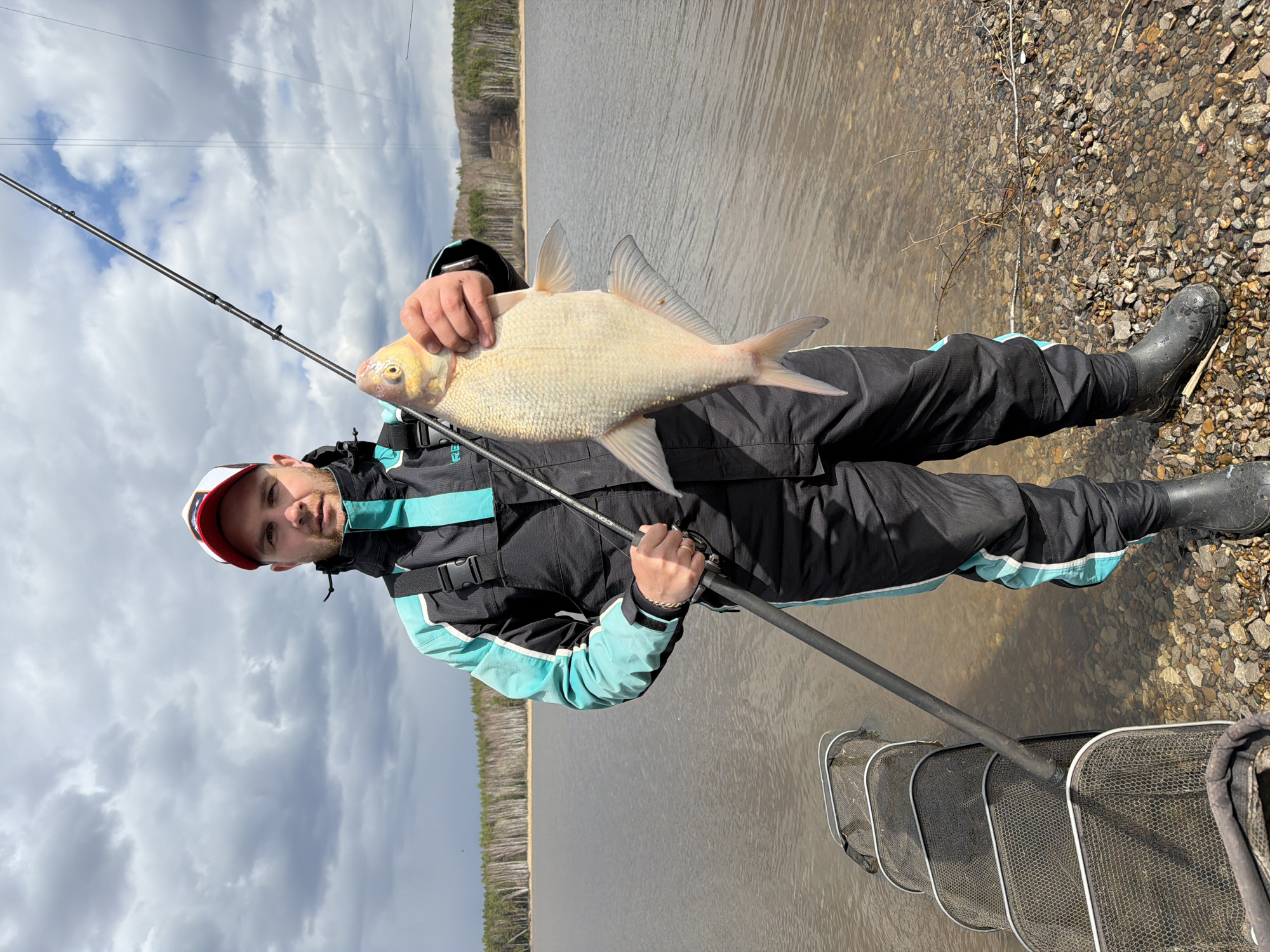 A man, dressed in black and turquoise fishing attire including a waterproof jacket, pants, and black rubber boots, stands on a rocky shore next to a body of water. He wears a red and white cap. He is holding a large, light-colored fish, possibly a bream or similar carp species, with both hands, showcasing his catch. The fish has silvery scales and a distinctive yellowish head.
In his right hand, the man holds a black fishing rod, on which the text "ICON" is faintly visible. To his right, a multi-section fishing net, likely a keepnet or landing net, is partially submerged in the calm, brownish-grey water.
The background features the expansive body of water, which appears to be a lake or a wide river, stretching towards a distant shoreline. A dense forest of coniferous trees lines the far bank. The sky is overcast with grey and white clouds, suggesting a cloudy daytime setting. The scene captures a moment of pride after a successful fishing outing.