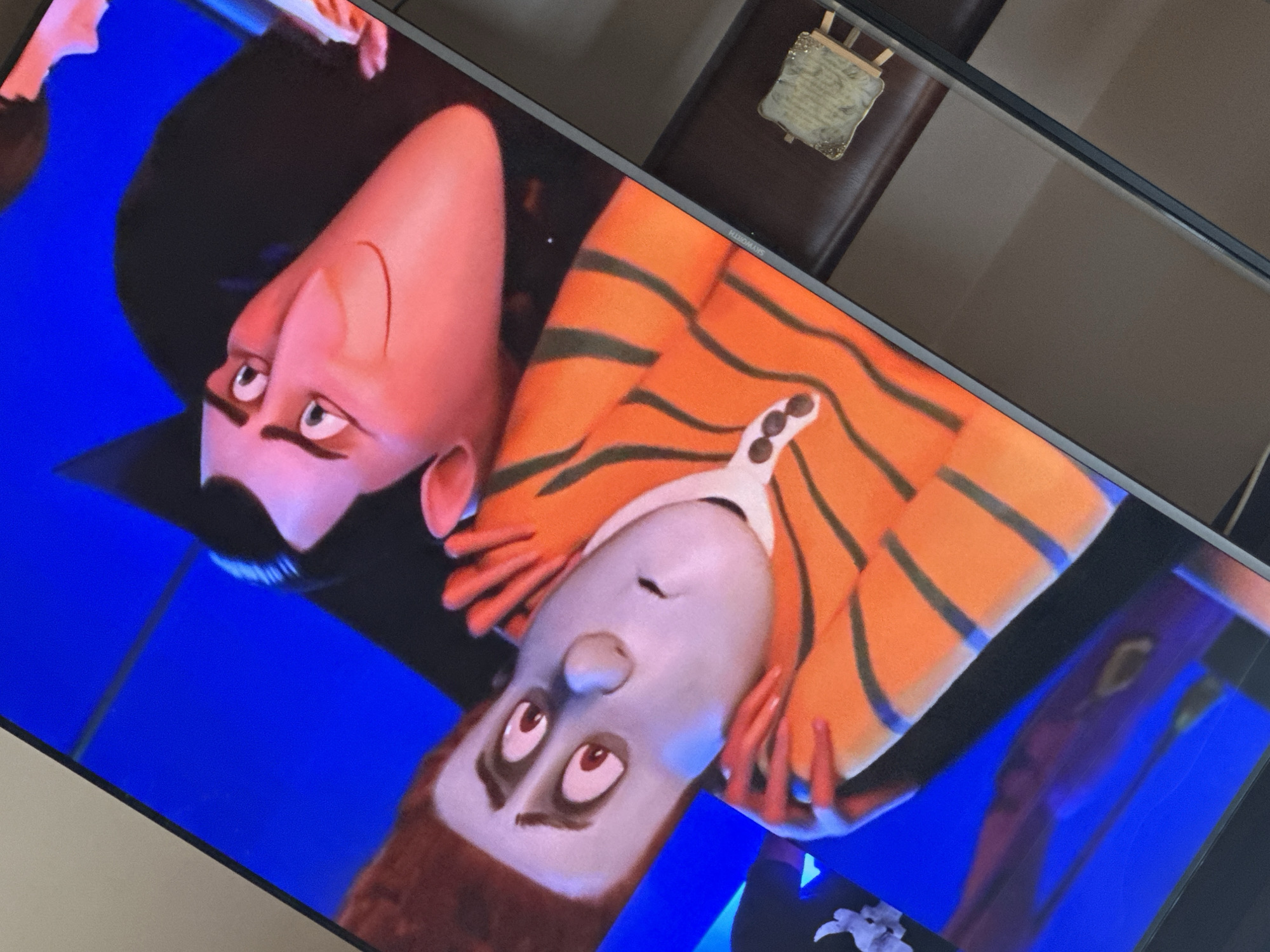 The image is a tilted close-up of a television screen showing a scene from the animated movie "Hotel Transylvania." Two characters are prominently featured: Dracula, with his signature slicked-back black hair and formal attire, and another character wearing an orange and black striped shirt. The background is a solid blue color. The photo was taken indoors. A dark chair with an ornate decoration on the back is visible in the upper right corner of the frame. The image's location is unknown.