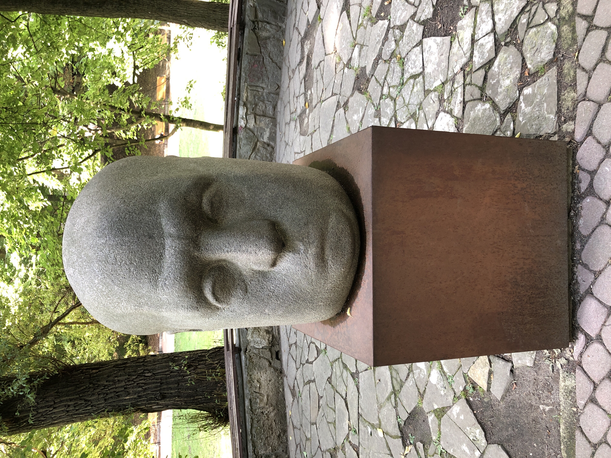 The image shows a large, gray stone sculpture of a human head mounted on a square, rusty metal pedestal. The sculpture is located outdoors, in what appears to be a park or garden. The ground surrounding the sculpture is paved with irregularly shaped stones, and there are trees and foliage in the background. Behind the sculpture and pedestal, there is a stone wall and a bench. The overall scene is tranquil and artistic. Based on the sculpture and its setting, this is likely the "Head of a Man" sculpture located in Stryiskyi Park in Lviv, Ukraine.
