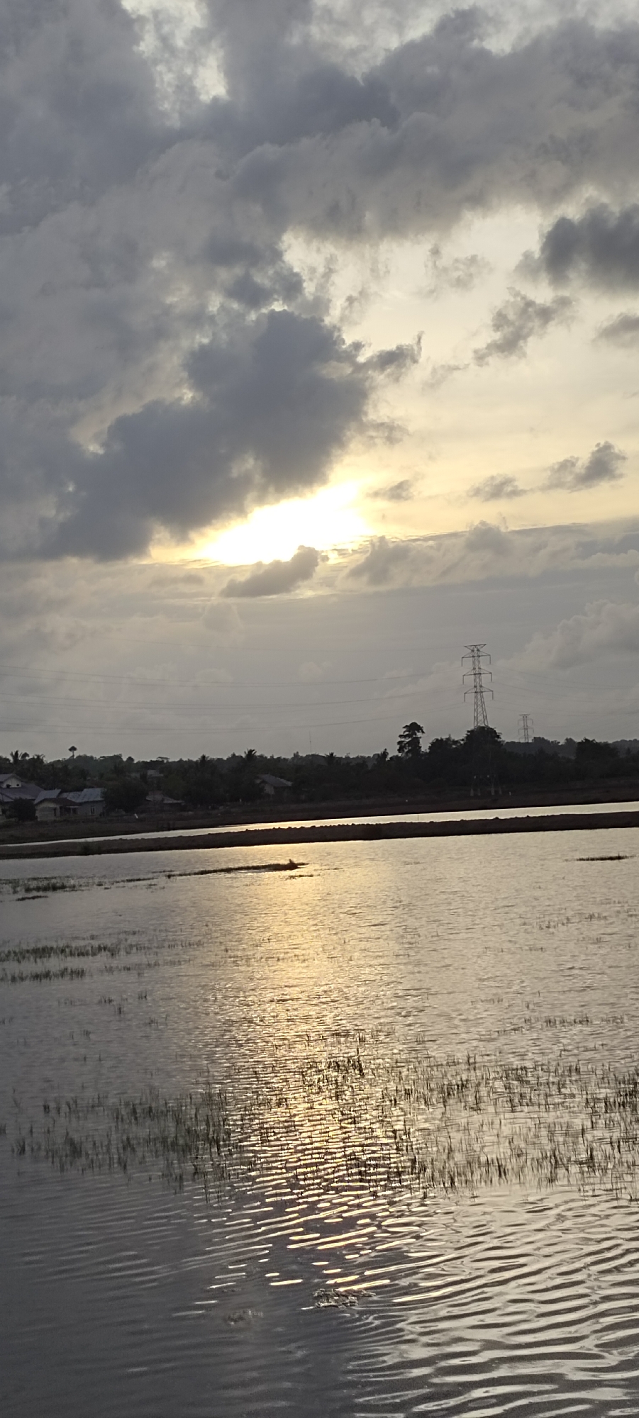 The image depicts a landscape at dusk or dawn in the city of Lhokseumawe, Indonesia. The foreground is dominated by a body of water, likely a pond or flooded field, with the surface reflecting the sunlight breaking through a layer of clouds. Tall grasses or aquatic plants are sparsely growing out of the water.
In the midground, a strip of land separates the water from the background. This land has some vegetation, and some manmade structures that look like homes. Several electrical transmission towers are also visible in the background.
The sky is overcast with thick, grey clouds, but a bright patch of sunlight illuminates the scene.