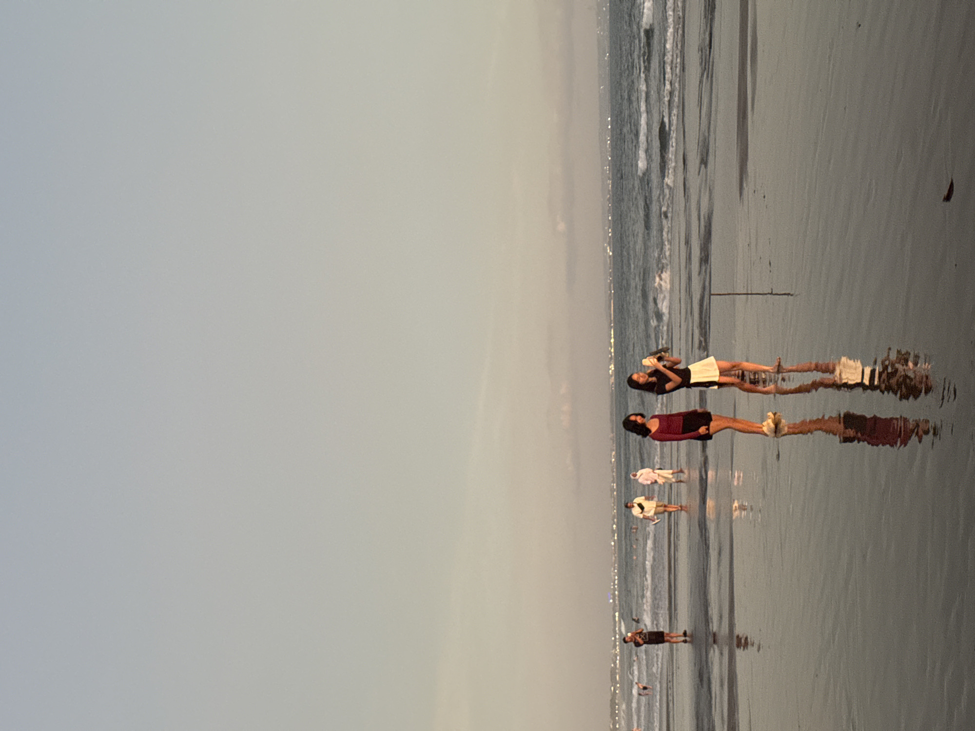 The image depicts a beach scene with several people. The setting appears to be a tropical beach, possibly in North Kuta, Indonesia, given the presence of a calm ocean with gentle waves and wet sand reflecting the sky. The time of day seems to be late afternoon or early evening, as indicated by the soft, diffused light and the long shadows cast by the people. The sky is overcast with a pale, muted gray hue, suggesting possibly cloudy weather or the approach of dusk.
The subjects are primarily people, with at least six individuals visible. Two women are most prominent in the foreground. One is lying down on the sand, holding a phone to take a picture. The other woman, standing, is wearing a burgundy top and dark shorts. Further down the beach, there are several other figures, some walking and some standing. One person appears to be wearing a white outfit, and another is in light-colored clothing. In the distance, more figures are visible, along with what looks like distant city lights or structures along the coastline.
The overall mood of the image is peaceful and relaxed, typical of a beach outing. The reflections on the wet sand add an artistic quality to the scene, blurring the lines between the figures and their mirrored images. There are no discernible objects other than the people and the natural elements of the beach. No text is visible in the image. The activity appears to be leisurely, with people enjoying the beach at their own pace.
