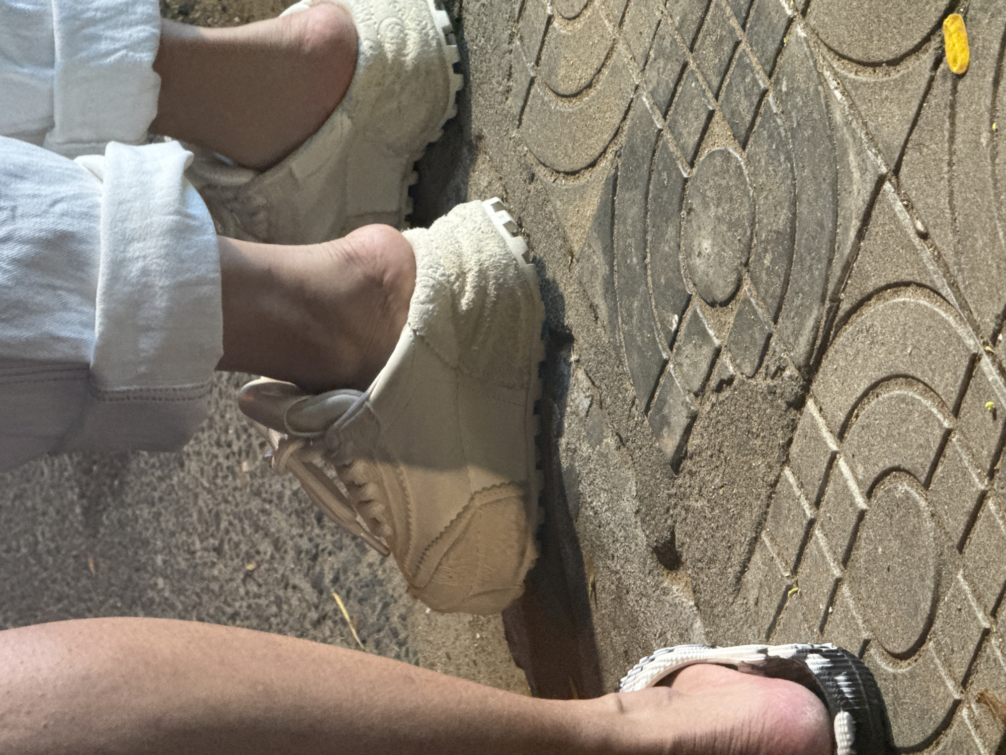 The image provides a top-down view of the lower legs and feet of at least two individuals standing or sitting on a decorative concrete pavement in Đà Nẵng, Vietnam.
On the left side, a person wears loose, white linen-like pants, rolled up at the ankles, revealing their bare lower legs and feet. They are wearing light, cream-colored slip-on shoes or slippers made of a soft, textured material, possibly faux fur or chenille, with a thick, ridged white sole.
In the lower right corner, another person's bare leg and foot are visible. This individual is wearing a black and white patterned sandal or flip-flop on one foot, with a woven or braided strap design.
The ground is composed of grey concrete paving tiles featuring an intricate geometric pattern of circles, squares, and arcs. Fine sand and dirt have accumulated in the grooves of these patterns, indicating an outdoor, possibly urban or park setting. The scene is brightly lit by what appears to be strong overhead sunlight, suggesting it is daytime and sunny, casting subtle shadows and highlighting the textures of the shoes and pavement. There is no visible text in the image.