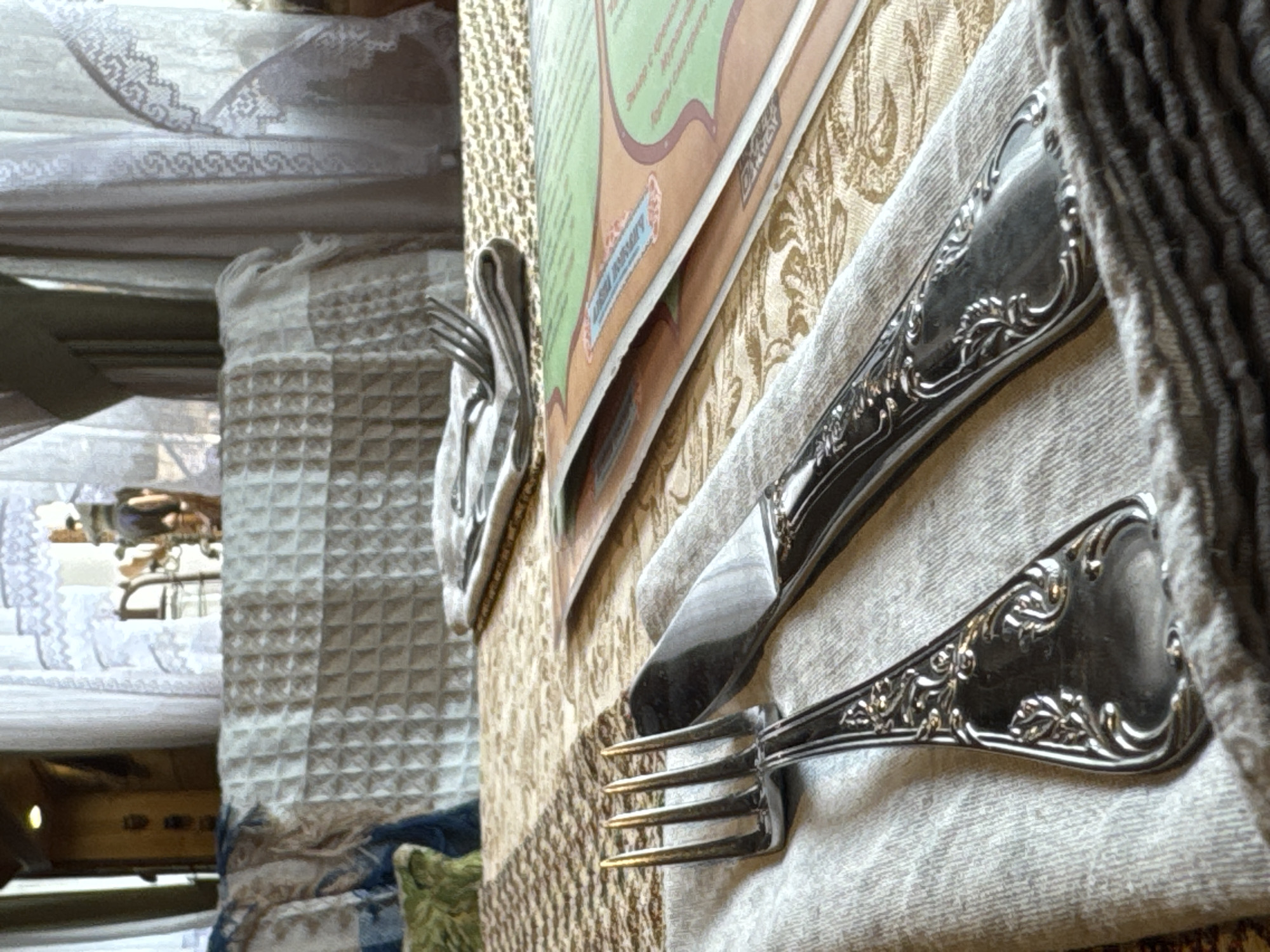 This is a close-up shot of a table setting, likely indoors. In the foreground, there is a decorative silver fork and knife arranged on a light-colored napkin. A folded menu or brochure is placed near the cutlery. The tablecloth has a patterned texture. In the blurred background, elements of a room are visible, including a chair and what appears to be a white curtain or bedspread with lace detail. The image was taken in the city of Ново-Никольское, Russia.