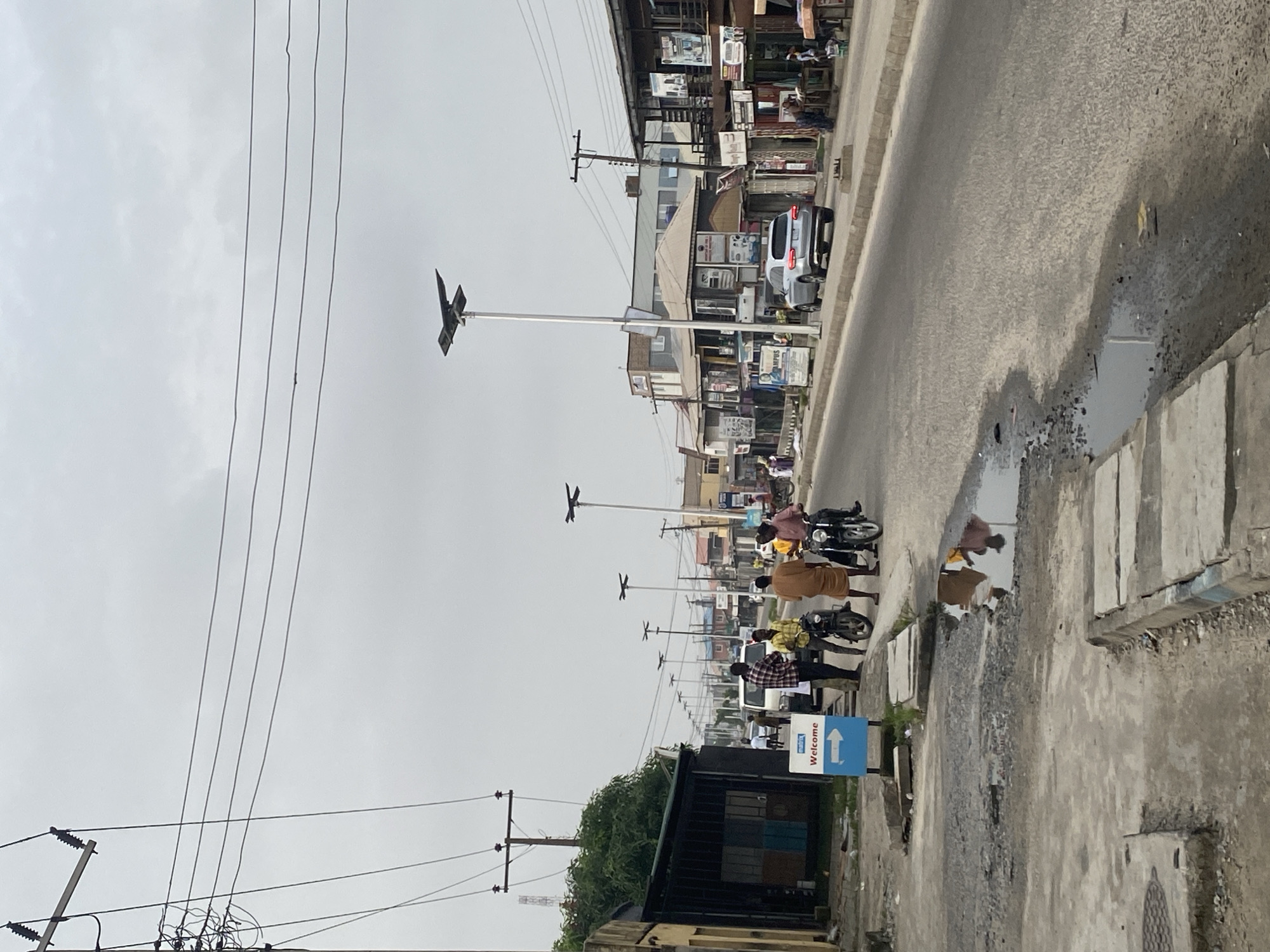 The image shows a street scene in Ado, Nigeria. A row of streetlights with distinctive cross-shaped fixtures line the road. Buildings flank the street, their facades displaying a variety of advertisements. Pedestrians, some on motorcycles, are present on the roadside. Overhead electrical wires are visible. On the right side of the road, there is a puddle of water reflecting the surroundings. A "Welcome" sign is planted nearby. The weather appears overcast.