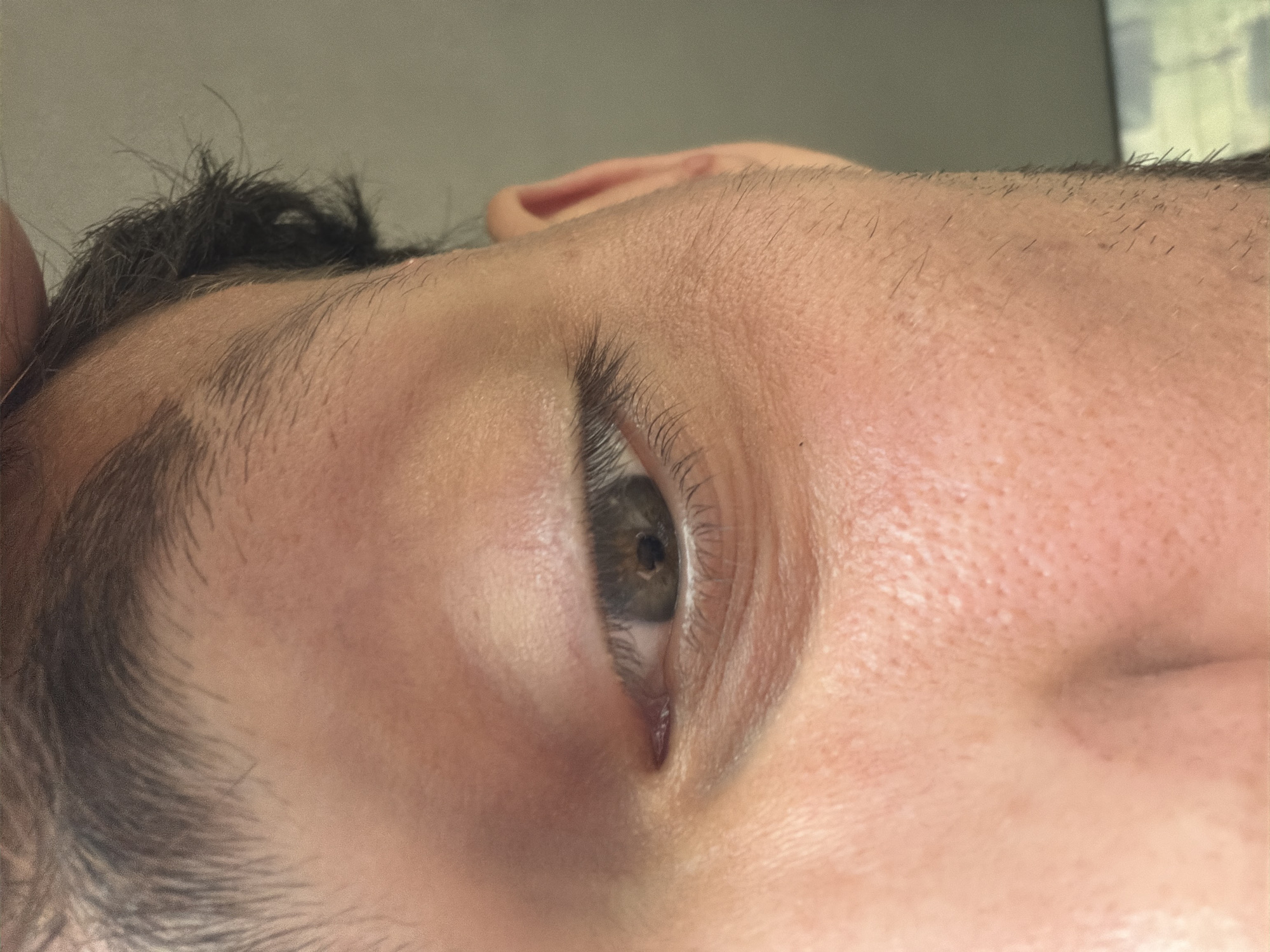 A detailed close-up image focuses on the right side of a human face, encompassing the eye, portions of the forehead, temple, cheek, and the bridge of the nose. The subject is an individual with light-to-medium skin tone and dark hair, evidenced by the eyebrow, eyelashes, and visible hairline. The right eye features a dark brown iris and is partially open, with the pupil clearly visible. The gaze appears directed slightly downward and to the left. The skin exhibits visible pores and fine lines, particularly around the outer canthus of the eye. Sparse, dark stubble is visible on the temple and cheek area, suggesting a male subject or recent hair growth. The ear is partially visible in the upper right background. The background is uniformly blurred and light greyish-beige, offering no identifiable features or landmarks to determine a specific location within the city of Rezovo, Bulgaria, or any other geographic area. No specific actions or interactions are depicted beyond the subject's static gaze.