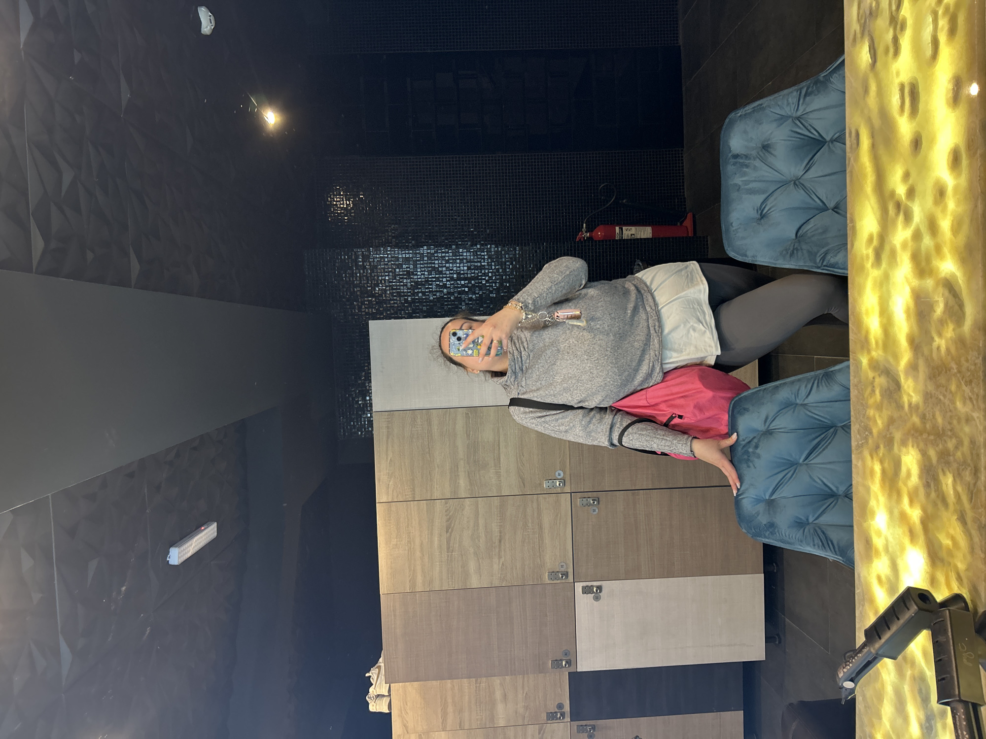 A woman stands in what appears to be a locker room taking a selfie in a mirror. She wears a gray long-sleeve shirt, white athletic shorts, gray leggings, and a pink bag slung over her shoulder. The lockers behind her are of varying shades of light brown and gray. A blue velvet chair is positioned to her right. The walls are decorated with black geometric tiles. A fire extinguisher is visible on the wall. Without additional information, it is not possible to confirm the location of this image in Plovdiv, Bulgaria.