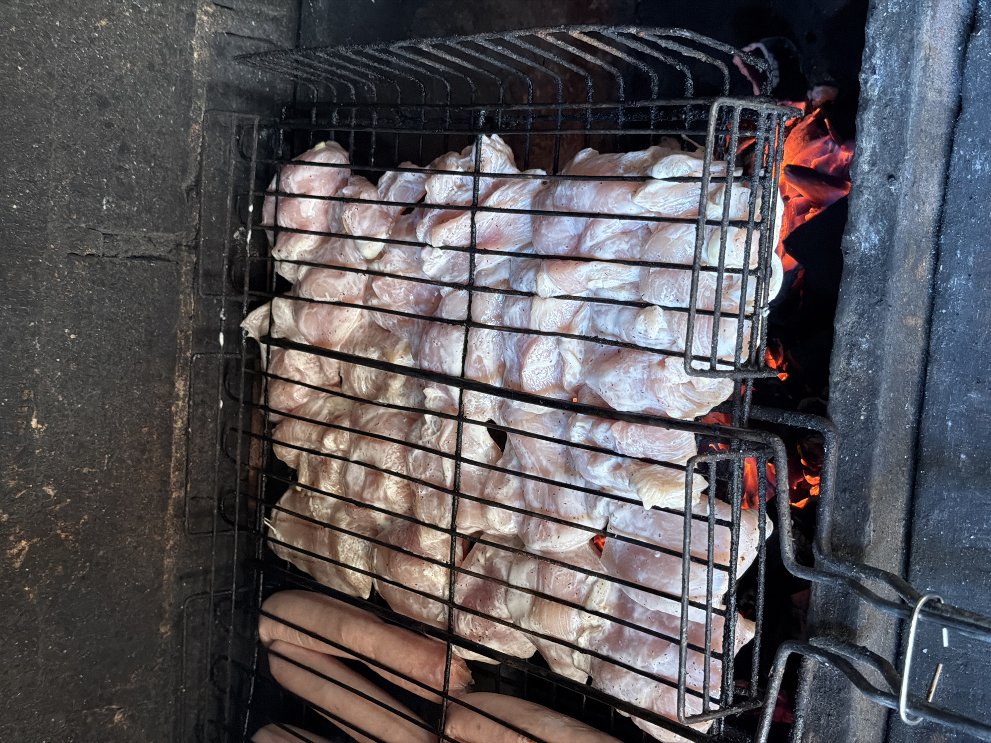The image displays a close-up, overhead view of food being grilled in an outdoor barbecue setup, situated in Chaikovsky, Russia. The primary subjects are numerous pieces of raw chicken, possibly cutlets or boneless thighs, coated in a white marinade and arranged densely within a black metal grilling basket or grate. Below these chicken pieces, several raw sausages are also visible, resting on a separate section of the grate.
The food is positioned over glowing red embers or coals, which provide the heat for cooking and are visible to the right and beneath the chicken. The grilling apparatus consists of dark, rough-textured stone or brick walls, suggesting a permanent, well-used outdoor grill or fireplace. The dark and somewhat sooty appearance of the structure hints at its frequent use.
The scene captures the initial phase of a grilling session, an activity commonly associated with outdoor gatherings. The overall lighting suggests it is daytime, but specific weather conditions are not discernible. There are no people visible in the frame, and no text is present in the image.