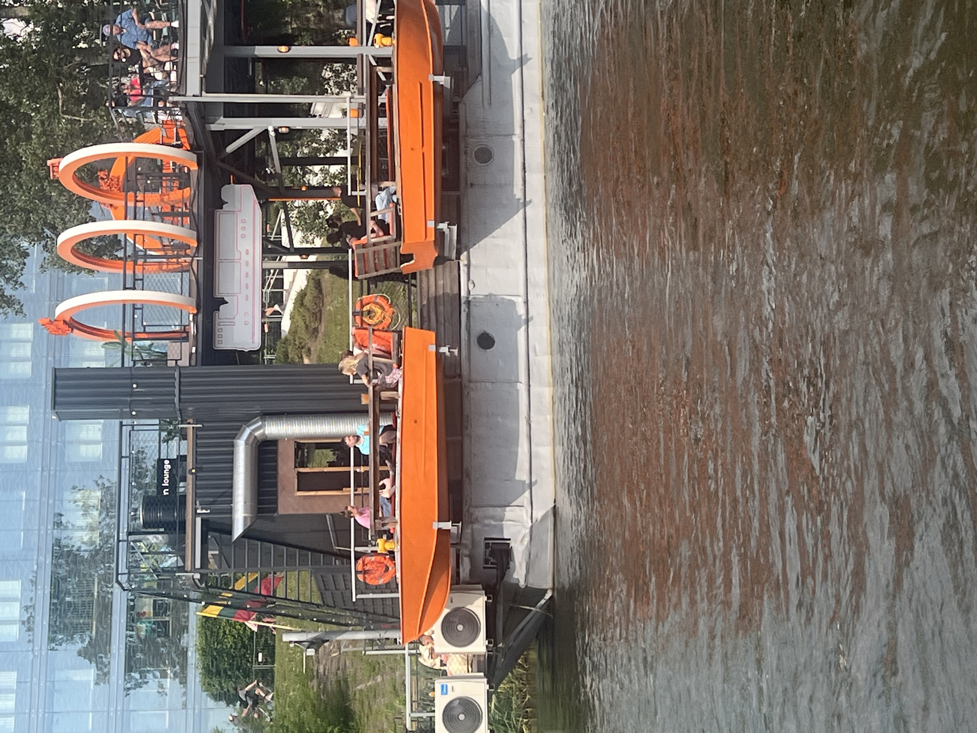 The image depicts a floating establishment on the Neris River in Vilnius, Lithuania. The structure appears to be a repurposed barge or pontoon, housing a multi-level lounge or restaurant area. The lower level features seating and tables where several patrons are visible, some appearing to be eating or drinking. Orange life rafts are affixed to the sides of the structure. Above this level, there are more seating areas, including a section with orange, arched structures that might be decorative or part of an attraction. A building with a glass facade, reflecting trees and sky, is visible in the background, suggesting an urban setting. The water in the foreground is choppy, indicating movement on the river. Two large air conditioning units are mounted on the side of the barge.