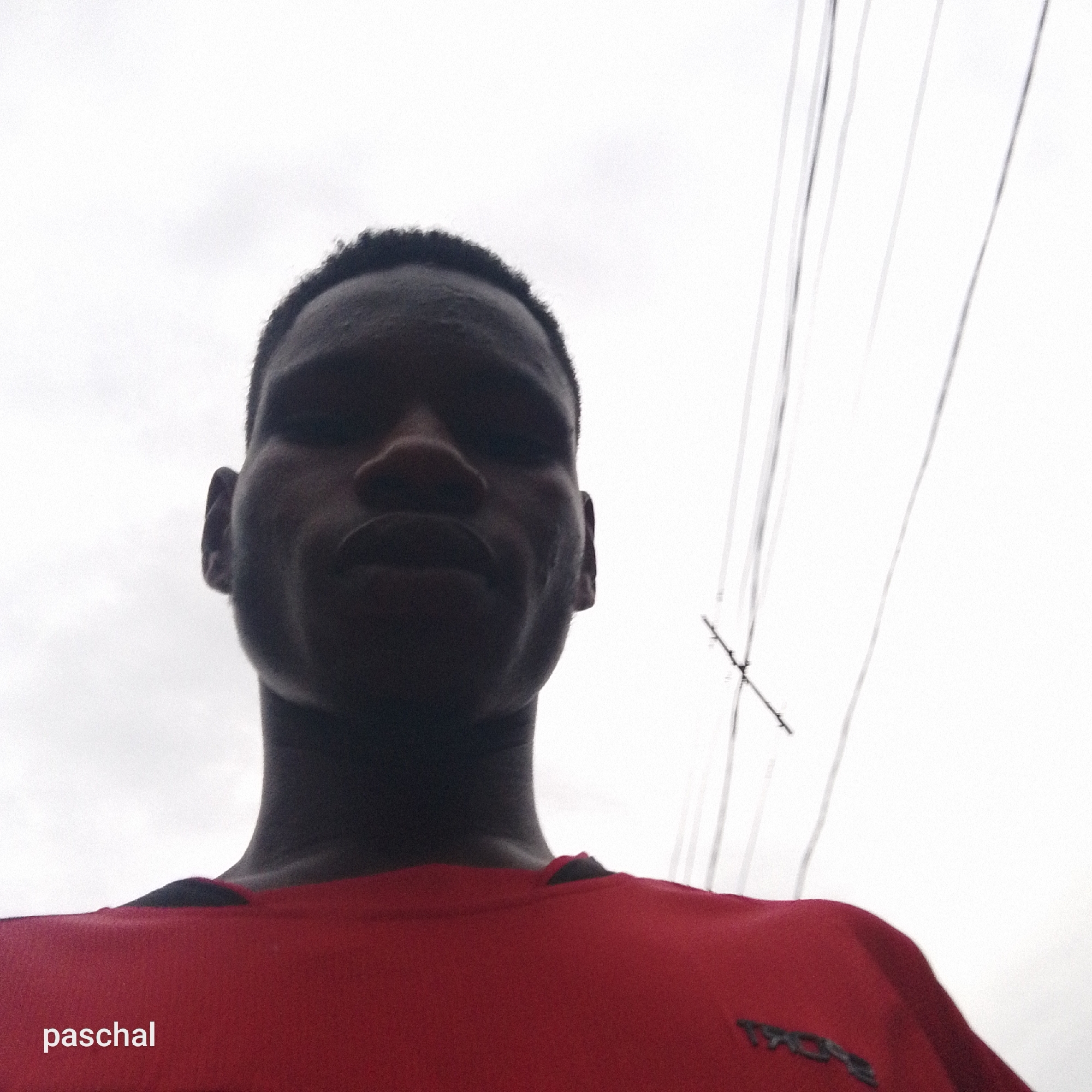 The image is a low-angle, head-and-shoulders shot of a young, dark-skinned man. He is looking directly at the camera, his face largely in shadow, giving him a neutral expression. He has short, dark hair. He is wearing a bright red t-shirt or jersey, and a blurry dark logo, possibly "TROX," is visible on his right shoulder area. In the bottom left corner, the name "paschal" is printed in white text.
The scene is outdoors, with an overcast or cloudy sky forming a bright, featureless background. In the upper right portion of the image, several thin, dark lines, resembling utility wires or cables, are visible, running diagonally across the sky with a cross-shaped junction point. The lighting suggests it is daytime, with diffused light from the cloudy conditions. No specific location landmarks are identifiable beyond the sky and utility lines. The man appears to be posing for a photograph, likely a selfie, given the perspective.