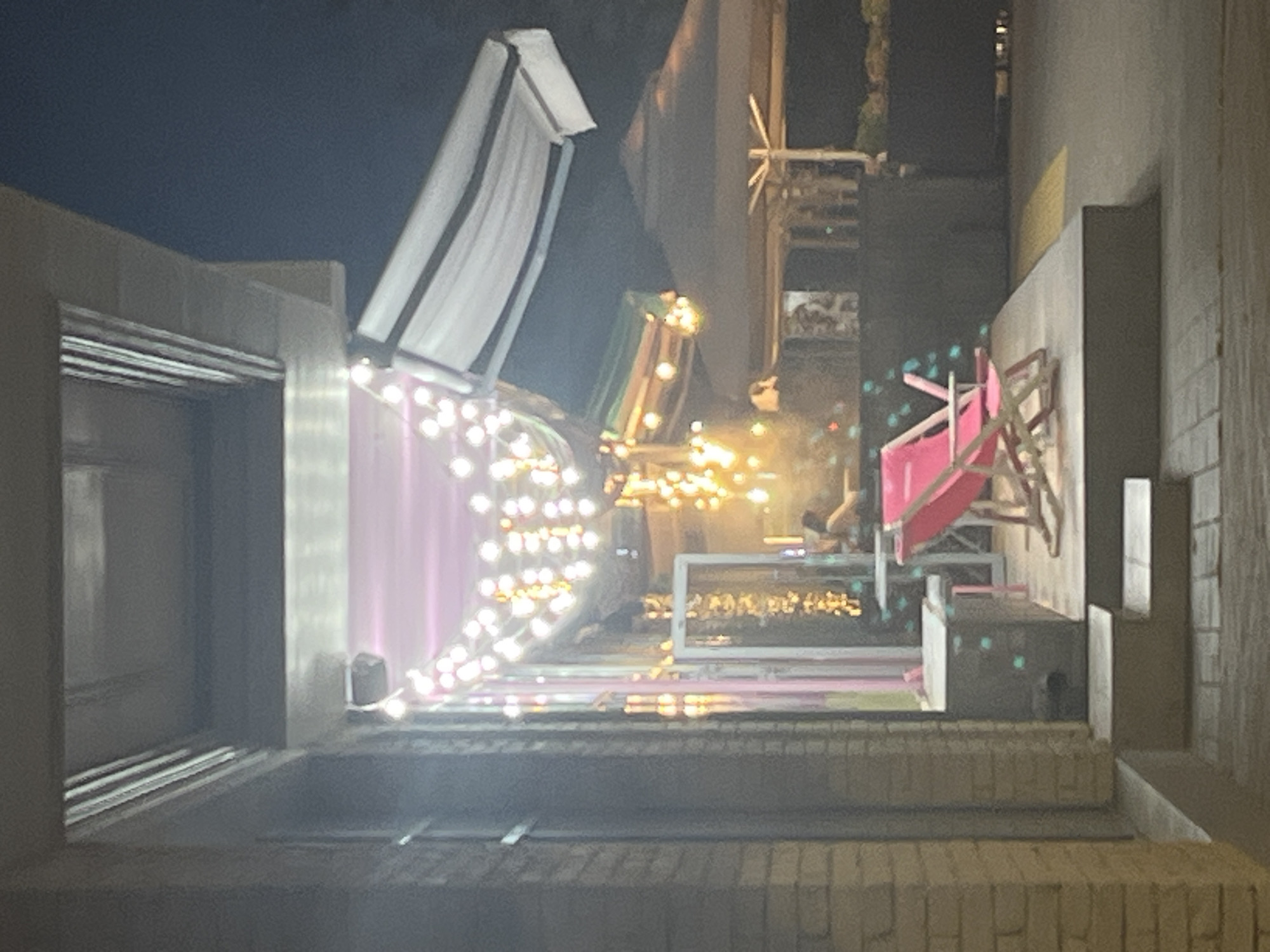 The image depicts an outdoor scene at night, possibly in Krasnodar, Russia, showcasing a creative display or installation. At the forefront, a textured brick surface forms a tiered base. Above this, a structure features a large, white, canopy-like element, tilted upwards, with dark trim. To its left, a vertical section of a building or wall is visible, with horizontal lines suggesting windows or architectural features.
Dominating the mid-ground is a vibrant display. A sheer, pink fabric hangs in graceful folds, illuminated by a dense string of warm white lights that cascade down it. Behind this, a multi-colored structure, possibly made of wood and metal, is partially visible, with some sections appearing green and others metallic. This structure is adorned with more warm string lights, creating a festive and inviting atmosphere.
Further to the right, a bright pink, angular object, resembling a geometric sculpture or a piece of furniture, is positioned. It has a star-like shape at its base and appears to be made of metal. In the background, more architectural elements and lights are visible, suggesting an urban or commercial setting. The overall mood is festive and illuminated, characteristic of a nighttime event or display. There is no visible text in the image.