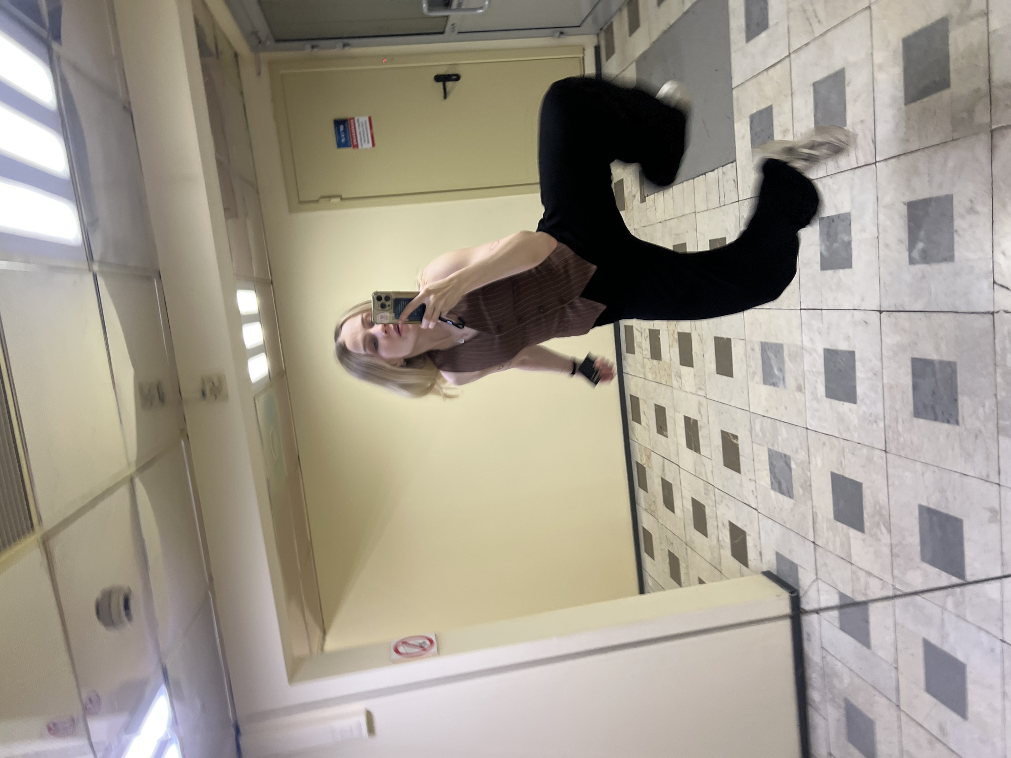 A woman is reflected in a mirror taking a photograph of herself with a mobile phone in what appears to be a hallway. The woman has blonde hair and is wearing a brown vest and black pants. The hallway has a beige wall with a door and a no-smoking sign. The floor is tiled with a pattern of squares. The photograph does not include enough information to determine if the location is Khimki, Russia.