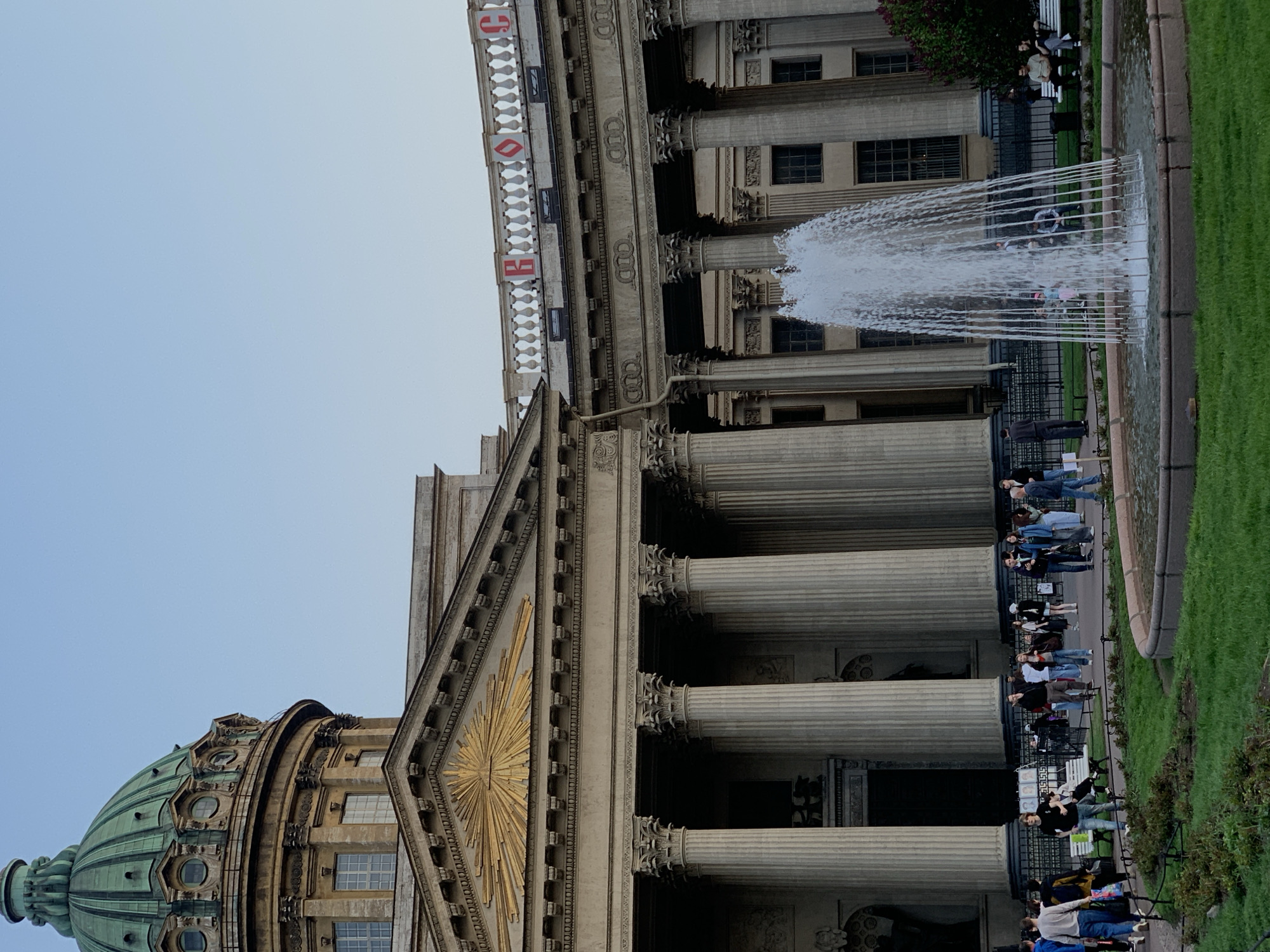 The image shows the Kazan Cathedral in Saint Petersburg, Russia, with its neoclassical architecture featuring colonnades, a dome, and a fountain in front. People are visible along the base of the building and near the fountain. The sky is overcast.