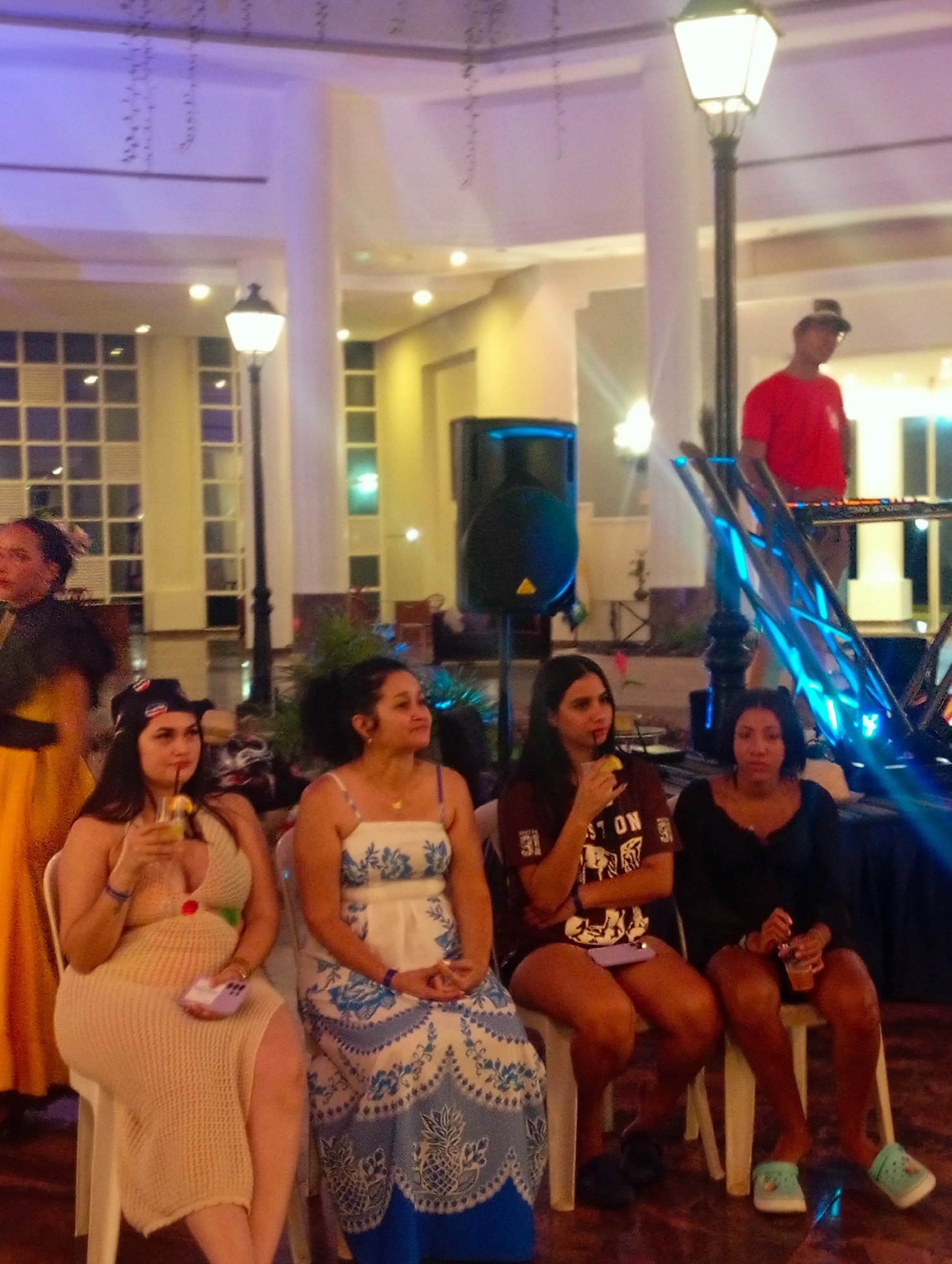 Here's a detailed description of the image:
**Setting:** The image appears to be taken at an indoor event or party, likely in the city of Pedro González, Venezuela. The interior has a high ceiling with decorative lights. There are large windows with multiple panes and pillars that are visible in the background.
**People:** Four women are seated on white plastic chairs in the foreground.
* Woman 1 (left): Wearing a beige crocheted dress and a hat, holding a drink in her hand.
* Woman 2: Wearing a blue and white patterned dress.
* Woman 3: Wearing a maroon t-shirt and black shorts, holding a drink and has a phone in her lap.
* Woman 4 (right): Wearing a black top and shorts and green croc shoes, holding a drink.
A DJ/musician wearing a red shirt and a hat is standing on a platform in the background, likely performing. There is another individual partially visible to the left of the woman with the hat.
**Objects:** The scene includes a black speaker, plants, and what appears to be a stage or platform for the DJ/musician.
**Actions:** The women in the foreground appear to be enjoying themselves, drinking and socializing. The DJ/musician is performing.
**Environment:** The ambiance appears to be relaxed and festive. There are string lights and other decorations visible in the background, suggesting a celebratory occasion.