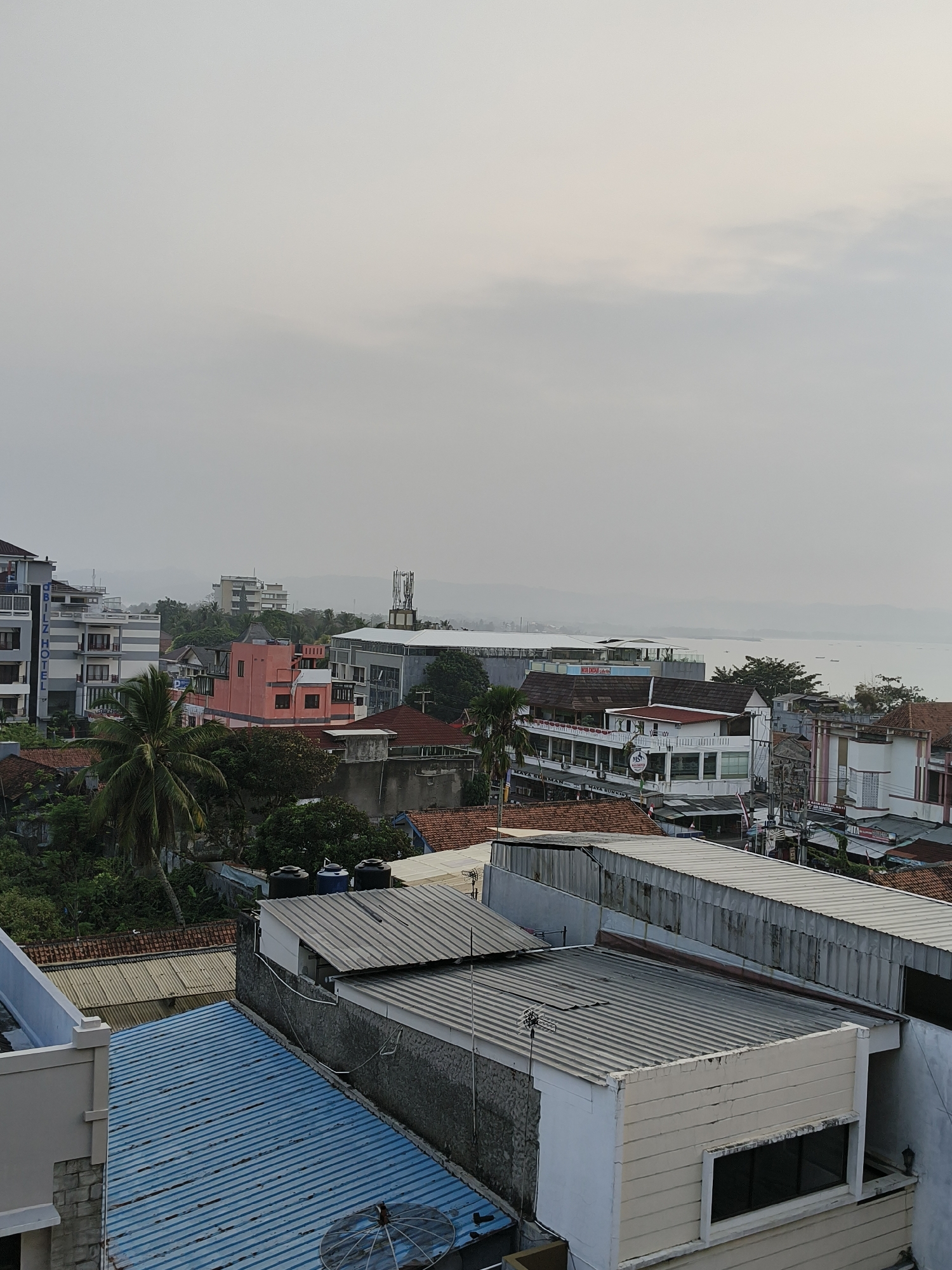 The image captures a coastal town with a dense collection of buildings and a body of water in the background. The architecture is varied, featuring multi-story buildings, some with balconies and signage, as well as lower-rise structures with tiled roofs. Vegetation, including palm trees and other greenery, is interspersed among the buildings. The sky is overcast, suggesting diffused daylight. The scene appears to be a view from an elevated position looking towards the sea. Signage on some buildings indicates commercial establishments, with one prominent sign reading "Q BILZ HOTEL." The overall impression is that of a developed coastal area, likely a tourist destination, given the presence of hotels and the proximity to the water. The location is identified as Desa Pagandaran, Indonesia.