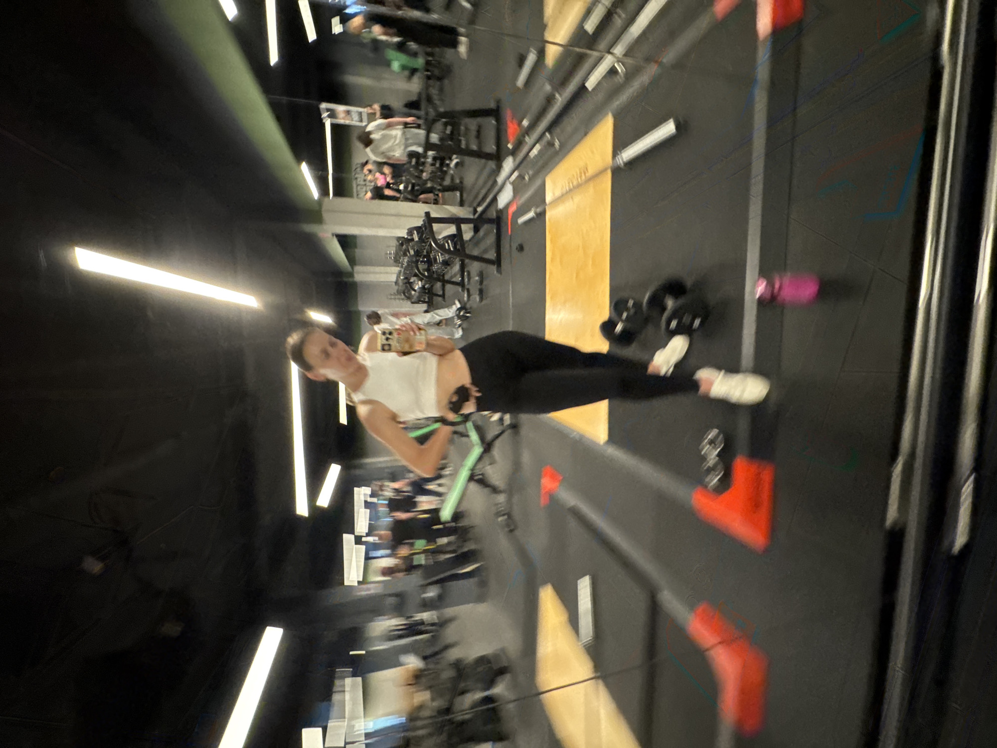 In this image, a young woman with brown hair is captured in a gym, likely in Sochi, Russia. She is wearing a white crop top and black leggings, and appears to be holding a phone in her right hand, possibly taking a selfie or recording a video. Her expression is neutral, and the overall mood seems relaxed and casual.
The gym setting is evident with the presence of exercise equipment, including weight racks and dumbbells scattered on the floor. The floor is dark, with some brightly colored accents on the equipment. Overhead, fluorescent lights illuminate the space, suggesting it's likely daytime or a well-lit indoor environment. The background shows other gym-goers engaged in their workouts, some in the process of lifting weights and others moving around the gym.
Notable details include a bright pink water bottle lying on the floor near a pair of dumbbells, and green resistance bands. The gym appears to be modern and well-equipped, with a variety of machines and free weights. The image is taken from a slightly low angle, emphasizing the subject and her immediate surroundings. There is no visible text in the image.
