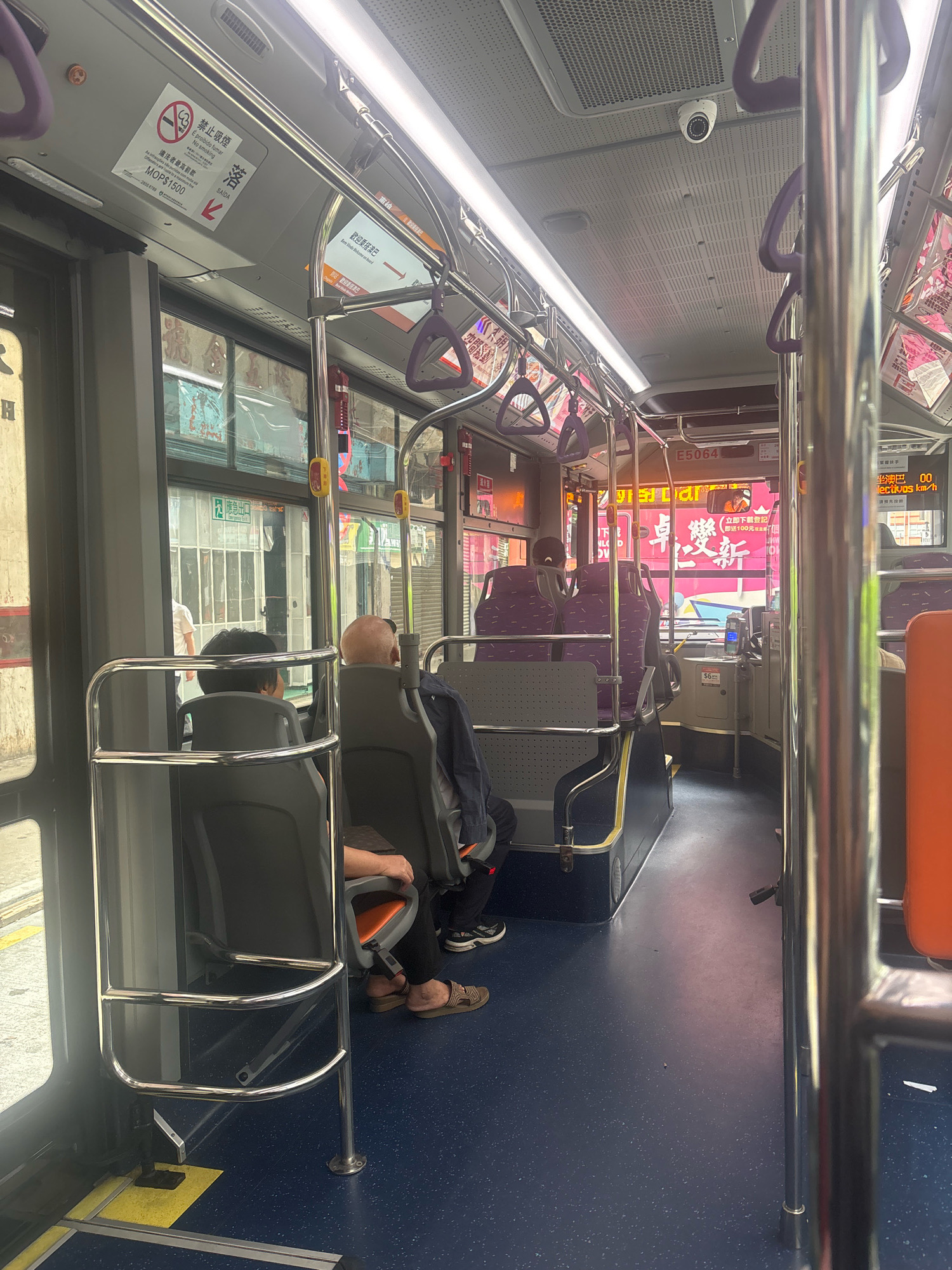 The image shows the interior of a public bus in Macau, China. The camera is positioned looking forward along the aisle. Several passengers are seated, including a man with a bald head in the middle row and a person with dark hair in the front row. The bus has purple fabric seats and metal poles for passengers to hold onto. Signs with Chinese and Portuguese text are visible on the interior walls, indicating regulations and information relevant to Macau. Outside the bus windows, buildings and street scenes of Macau are visible. A security camera is mounted on the ceiling of the bus. The floor of the bus is dark blue with yellow markings.