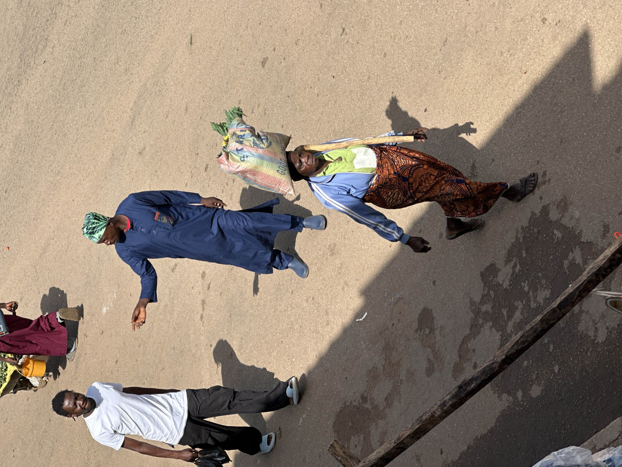 In Bukuru, Nigeria, a group of people are walking on a paved street. In the foreground, a woman with dark skin wears a blue jacket over a light green shirt and an orange and blue patterned wrap skirt. She has a large, colorful woven bag filled with green vegetables balanced on her head and carries a wooden pole over her right shoulder. To her left and slightly behind her, a man in a blue tunic and a green and white patterned headwrap gestures with his right hand. He is walking towards a man in a white t-shirt and black pants, who is walking away from the camera and holding two black bags in his left hand. In the background, partially visible, are two other people. One person is wearing a maroon garment and pushing a cart filled with yellow buckets and green produce. The other person is wearing a dark garment. The ground is a light brown asphalt surface with some wet patches and shadows from the people and a wooden beam in the lower left corner. The scene is illuminated by bright sunlight, casting distinct shadows.