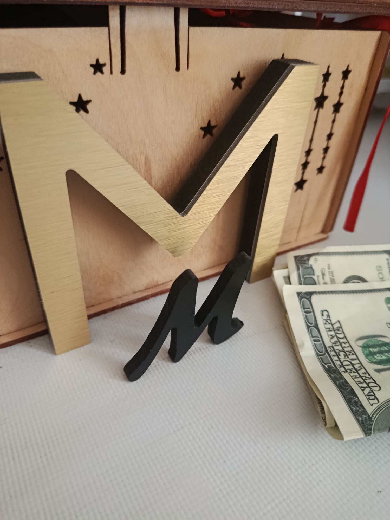 In an indoor setting with a white, textured surface, a wooden box with star cutouts and decorative lines is visible. A large, gold-colored letter "M" is attached to the front of the box, with a smaller, black, three-dimensional letter "m" positioned in front of it and slightly to the right. To the right of the letter "m", a stack of US hundred-dollar bills is fanned out. The scene is dimly lit, with the focus on the central objects. The background is blurred, suggesting a shallow depth of field.