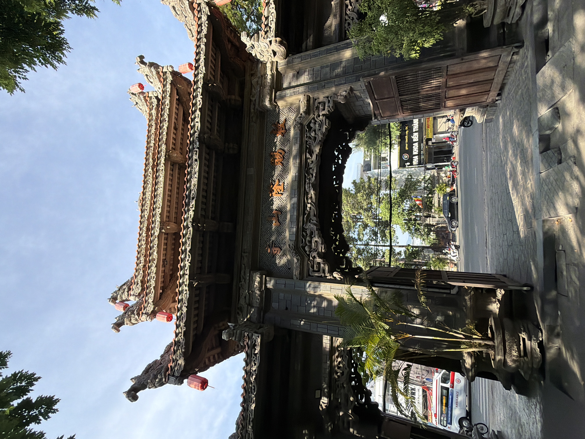 The image captures a detailed view of a traditional ornate wooden gate in Nha Trang, Vietnam, under a bright, partly cloudy sky. The gate is intricately carved with dragon-like figures and decorated with red lanterns. The roof of the gate features layered tiles and ornate carvings. Through the archway, a street scene is visible, including an ambulance, motorcycles, and a black car. Buildings line the street, with signs suggesting businesses, including a dental clinic labeled "YOUR TEETH SAY OK." Lush green trees frame the scene, casting shadows on the paved walkway in the foreground. A potted palm tree stands to the left of the gate. The overall impression is of a culturally rich and active urban environment.