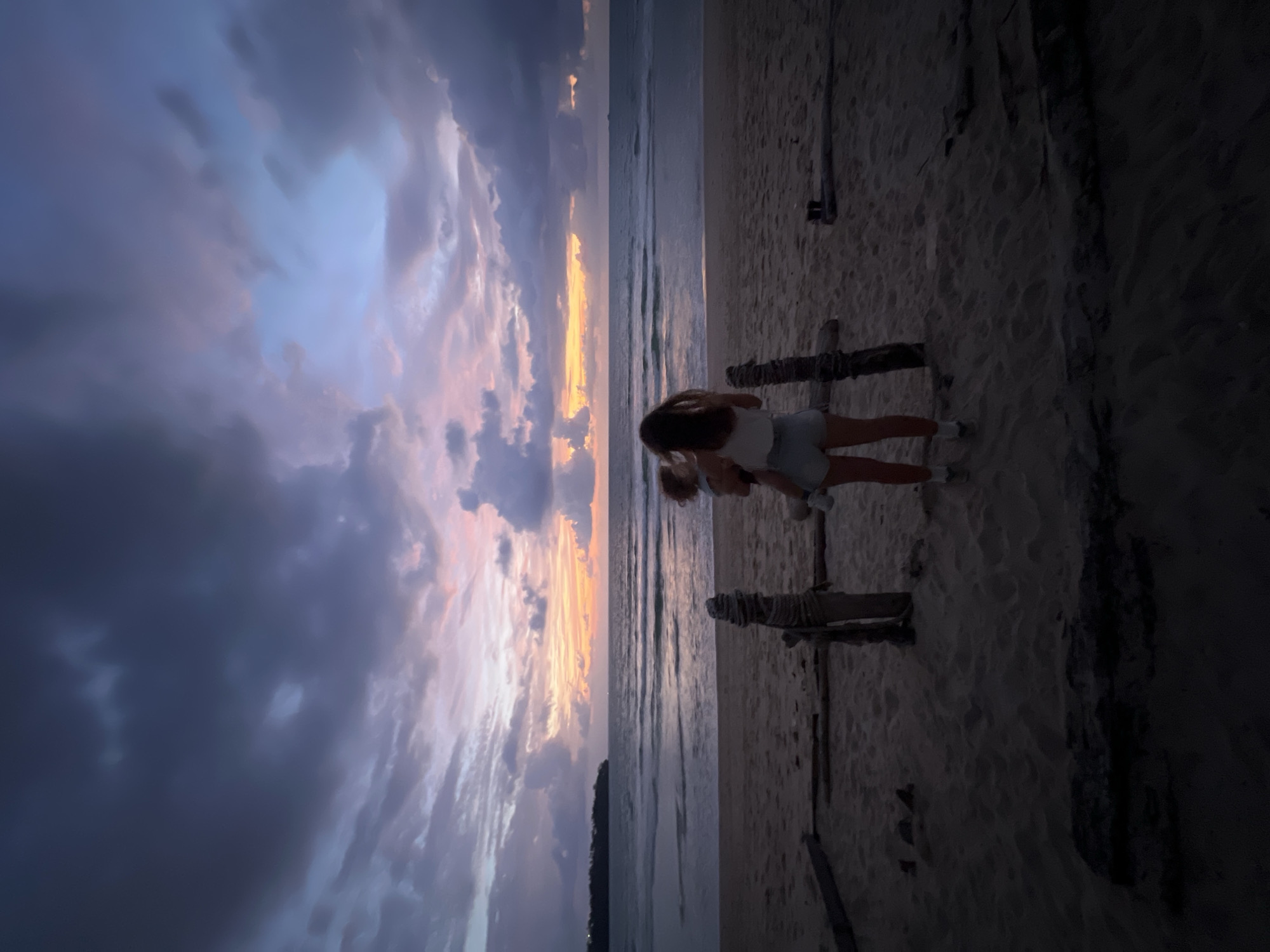 A full shot captures a woman holding a child on a beach at dusk in Choeng Thale, Thailand. The woman, viewed from behind, wears a white tank top and denim shorts. The child, also facing away, is held in her arms. They stand on a sandy beach with several wooden posts and logs scattered across the foreground, suggesting remnants of a structure. The ocean stretches out behind them, with gentle waves rolling onto the shore. The horizon is marked by a distant landmass to the left and a faint ship to the right. Above, a dramatic sky displays a gradient of colors, transitioning from deep purples and grays in the upper portions to softer pinks, oranges, and yellows near the horizon, indicative of a sunset or twilight. The overall scene evokes a tranquil and serene atmosphere.