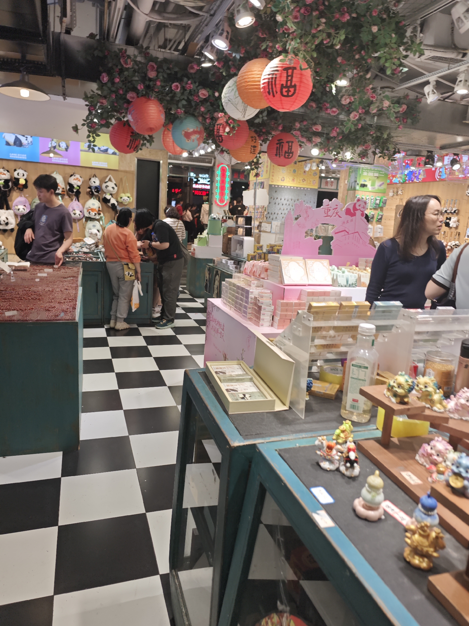 The image depicts the interior of a retail store in the Yuzhong District of China. The floor is covered with black and white square tiles. Several customers are present, interacting with the merchandise. A man in a gray shirt stands behind a counter, attending to customers. Other individuals are examining goods displayed on tables and shelves. The store's decor includes hanging red lanterns with Chinese characters and floral arrangements. Products are arranged on the shelves, including small figurines and gift items. A woman in a dark blue shirt appears to be observing the scene.