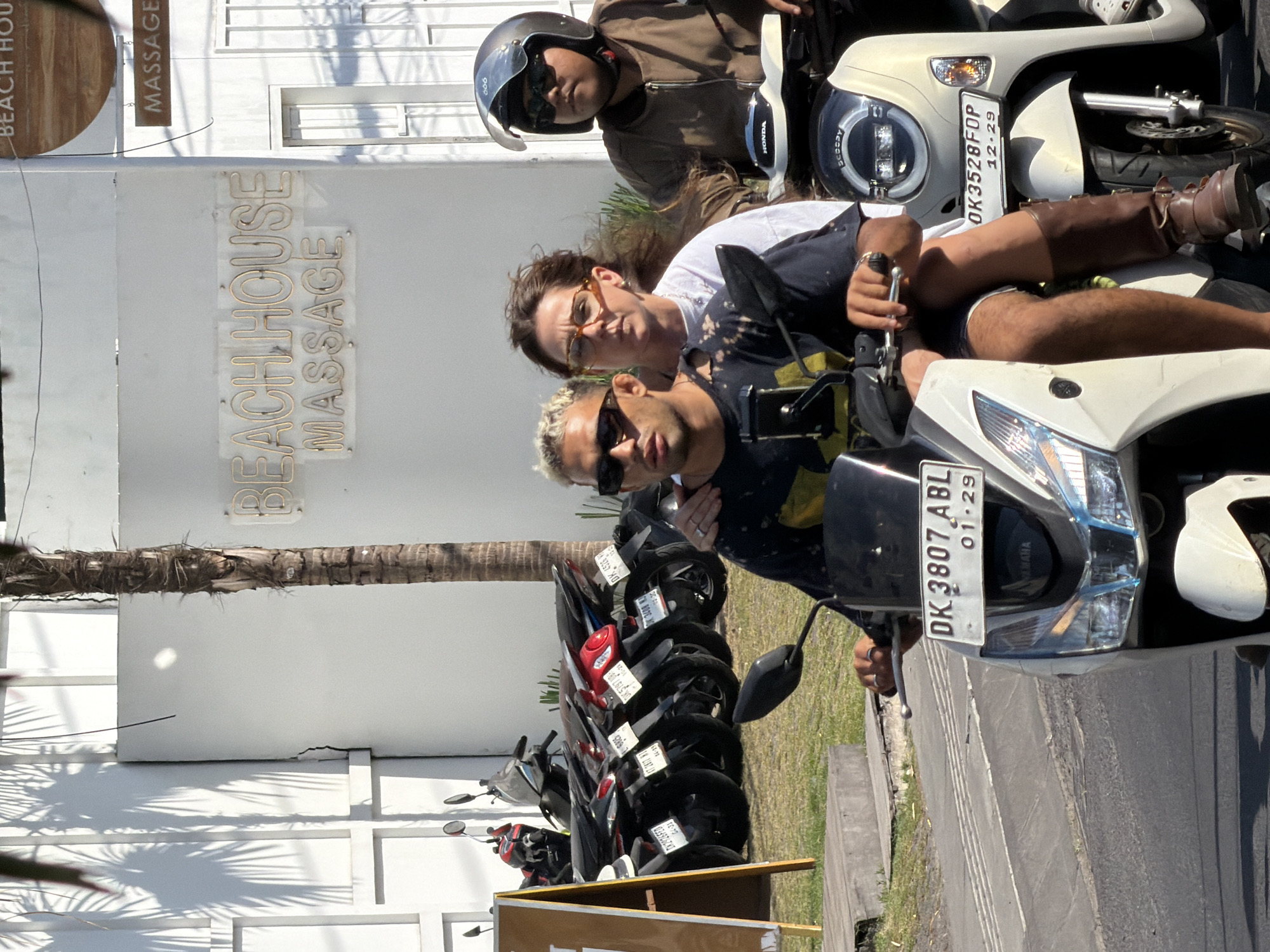 The image shows three people riding on two white scooters in front of a building with a "BEACH HOUSE MASSAGE" sign. The first scooter, in the foreground, carries two people. The person in the front, a man with blond hair, sunglasses, and a dark t-shirt with a tie-dye pattern, is driving. He is wearing dark shorts. A woman with brown hair and glasses, wearing a white t-shirt, is riding as a passenger behind him. She has one arm around the driver's shoulders. The second scooter, to the right and slightly behind, is driven by a man wearing a helmet and sunglasses, and a brown jacket. He appears to be of Asian descent. The scooters are parked next to a row of other scooters. The environment appears to be a sunny day in a tropical location, possibly a tourist area in North Kuta, Indonesia, given the scooters and beach-themed signage. The building is white with large windows and palm tree shadows are cast on its wall.