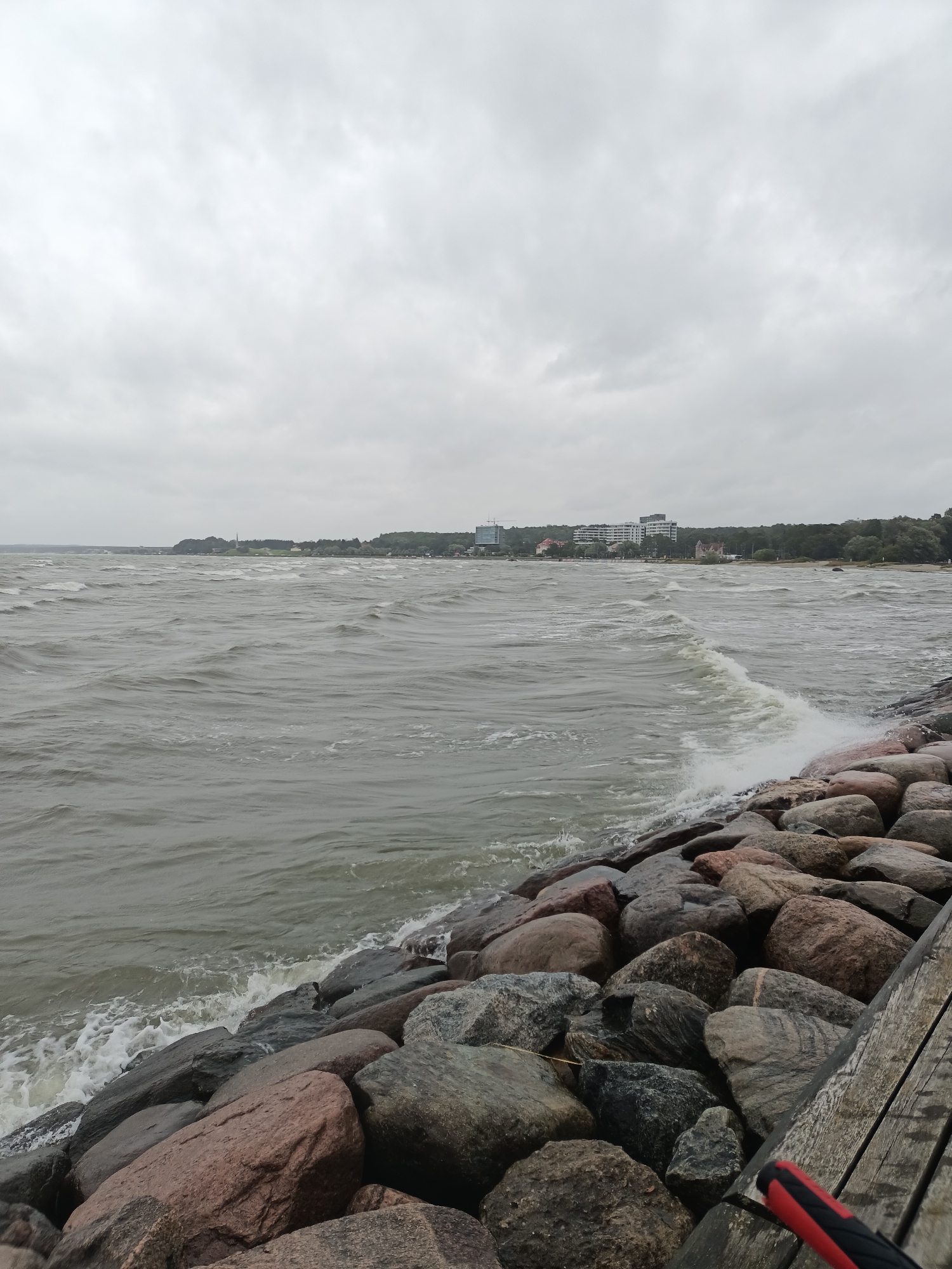 The image depicts a rough sea with choppy waves crashing against a rocky shore and a wooden pier. In the background, a city skyline with buildings and trees is visible. The sky is overcast and grey, contributing to a somber atmosphere. The scene appears to be a coastal area in Tallinn, Estonia, possibly near the Pirita promenade, known for its picturesque seaside views and breakwaters.