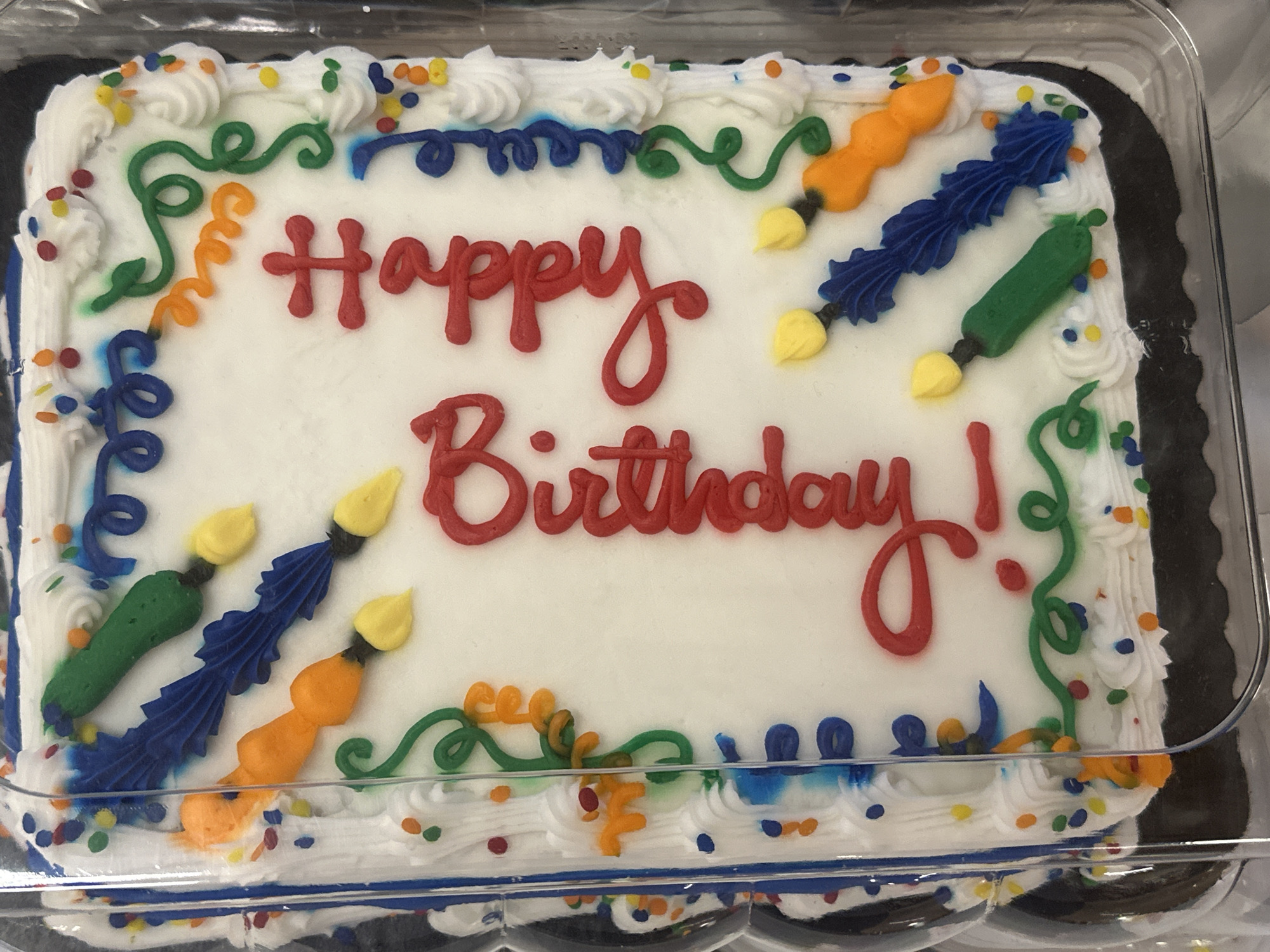 This is a top-down, slightly angled shot of a white sheet cake decorated with colorful frosting. The cake is in a clear plastic container. The frosting design features a red script spelling out "Happy Birthday" vertically on the right side of the cake. To the left of the text, there are several piped frosting decorations resembling paintbrushes with yellow tips. These brushes are in various colors, including green, blue, and orange. There are also swirls of blue and green frosting around the edges of the cake, interspersed with small colorful candy dots that resemble sprinkles. The cake itself appears to be on a black cake board. The image does not provide any context about its location within the city of Camp Hill, United States of America.