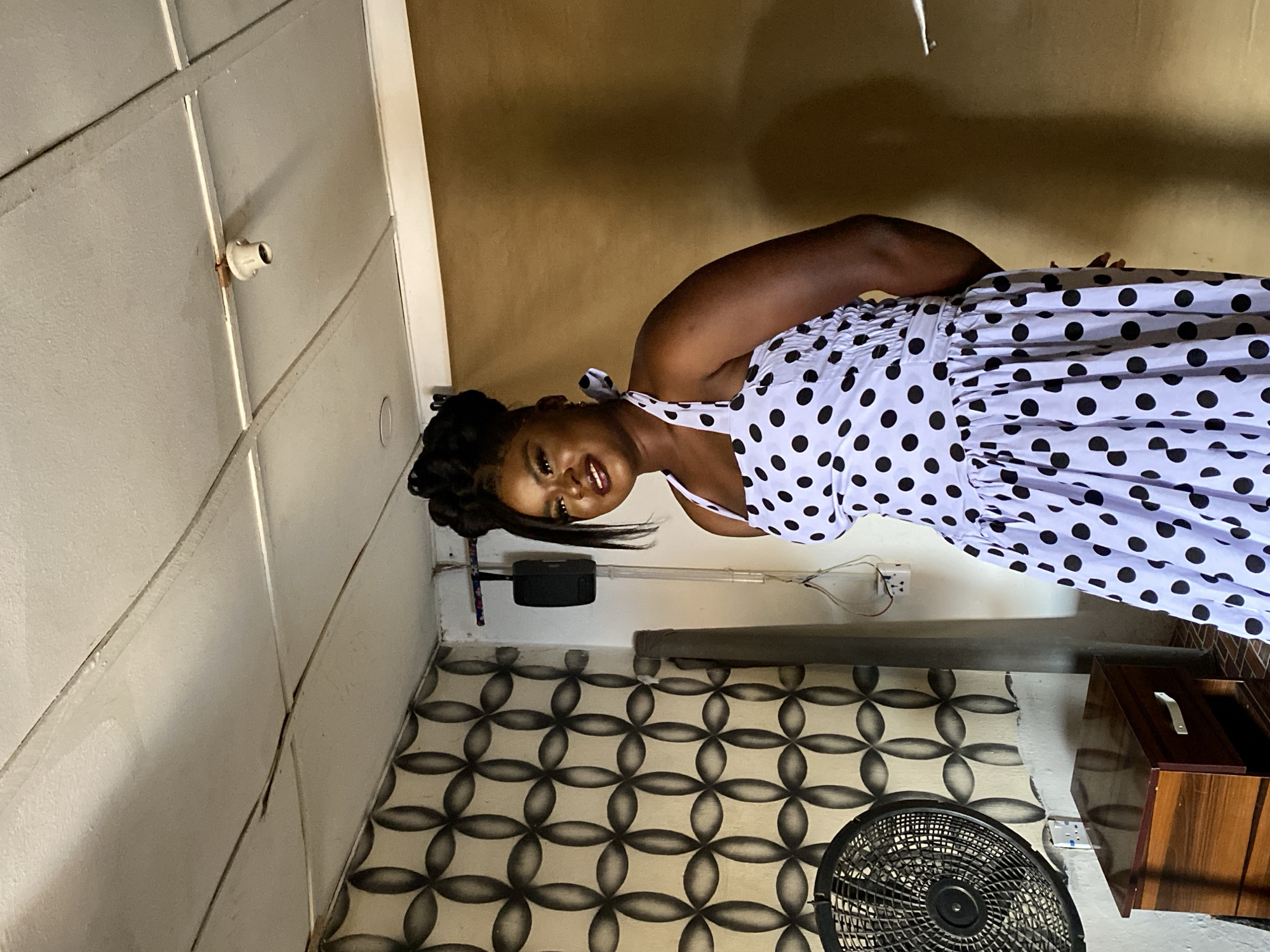 A portrait-oriented photo, taken indoors in Vom, Nigeria, shows a dark-skinned woman smiling brightly at the camera. She stands against a light-colored wall, wearing a white sleeveless dress adorned with large black polka dots. The dress features thin spaghetti straps and a gathered or smocked bodice, with a full, flowing skirt. Her dark hair is styled in an elaborate updo, possibly braids or twists, pulled back from her face.
The room has a utilitarian feel. The walls are light beige or off-white, with noticeable seams, possibly from panels or uneven plaster, and some discoloration. A white light bulb holder, showing some rust, is mounted on the upper wall. The top portion of the wall beyond the woman's head is painted a muted yellow or beige.
The floor is covered with patterned tiles featuring a repeating geometric design of interconnected circles or four-petal flowers in shades of light and dark gray or black. In the bottom left, a black cylindrical or rectangular speaker is mounted on the wall. Towards the bottom right, a black electrical fan is partially visible, and a dark wooden cabinet or drawer unit with a silver handle is also seen. White electrical outlets with visible wires are present on the wall, suggesting practical, functional wiring. The overall lighting suggests daytime. There is no visible text.