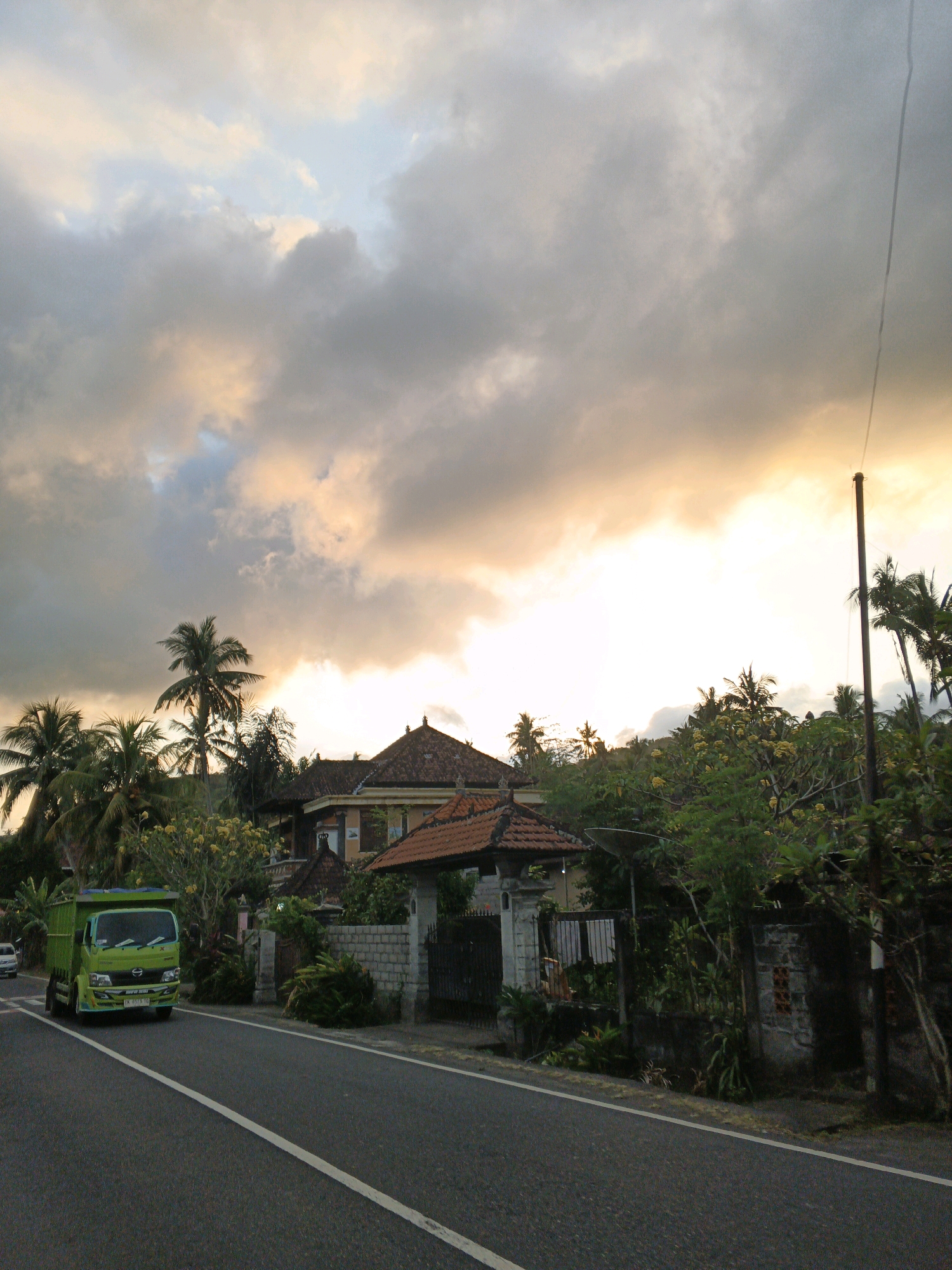 The image depicts a roadside scene in Manggis, Indonesia, during what appears to be either sunrise or sunset. The sky is filled with dramatic clouds, illuminated by the warm glow of the setting or rising sun. On the left side of the asphalt road, a bright green truck is traveling away from the viewer. The road is marked with a white line. On the right side of the road, there is lush green vegetation, including palm trees, and several traditional Balinese-style houses with dark tiled roofs. A stone wall and a gate are visible in front of one of the houses. A tall, slender utility pole stands to the right of the houses. The overall impression is one of a tropical rural or semi-rural environment bathed in soft, atmospheric light.