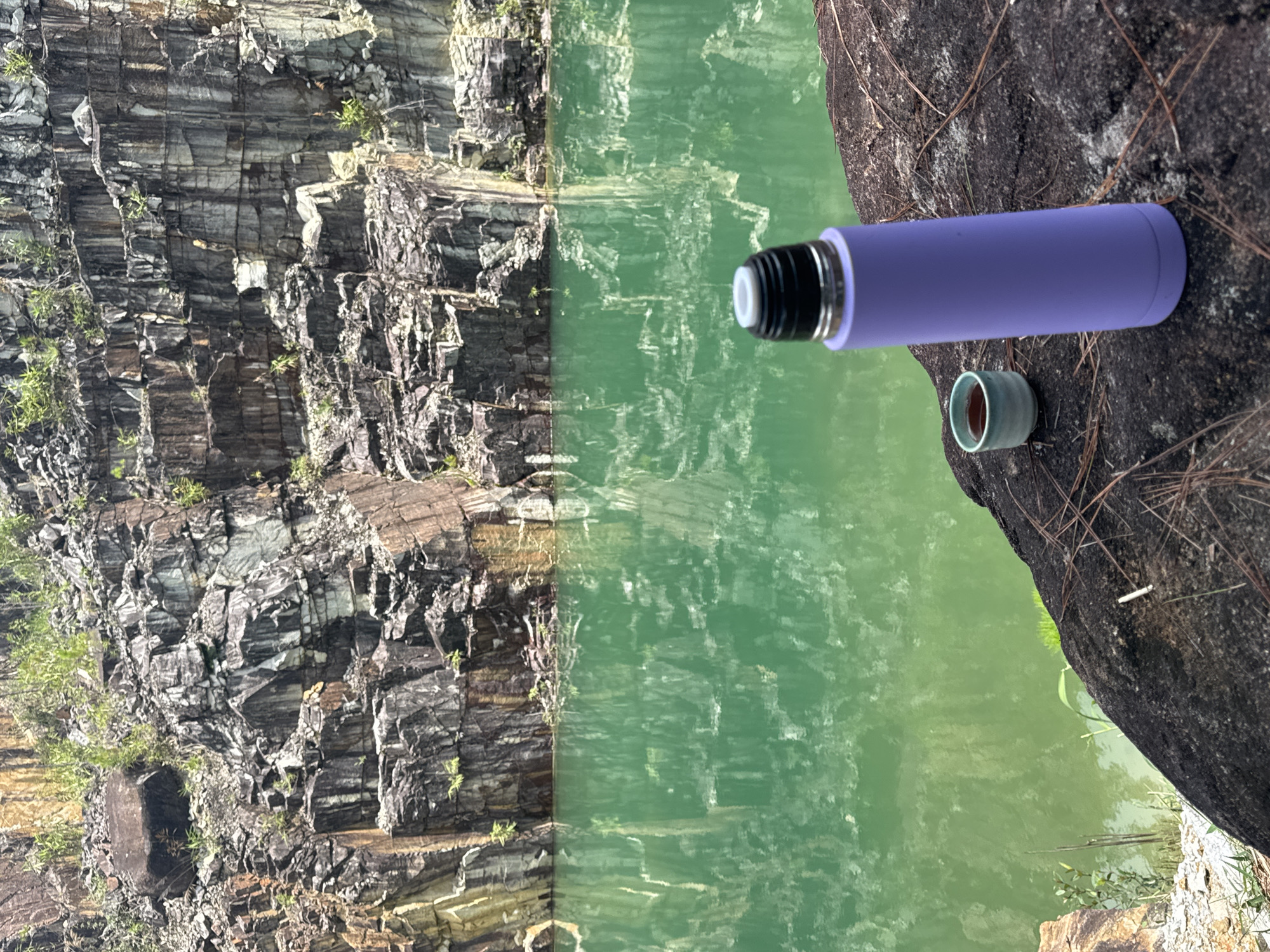 An environmental scene captures a serene moment with a purple thermos and a small ceramic cup filled with a dark liquid resting on a large, dark rock in the foreground. Beyond the rock, a vast body of calm, emerald-green water reflects the imposing, rugged cliff face of a quarry. The quarry walls are characterized by distinct, layered rock formations with variations in color, ranging from dark gray and brown to patches of ochre and white. Sparse green vegetation clings to the rocky surfaces and lines the water's edge. The presence of pine needles scattered on the rock suggests a natural, possibly wooded, environment. The location is identified as a quarry in the Lang Biang area of Da Lat, Vietnam, a region known for its natural beauty and mountainous terrain. The overall impression is one of tranquility and natural splendor, with the man-made object of the thermos and cup juxtaposed against the raw, geological landscape.