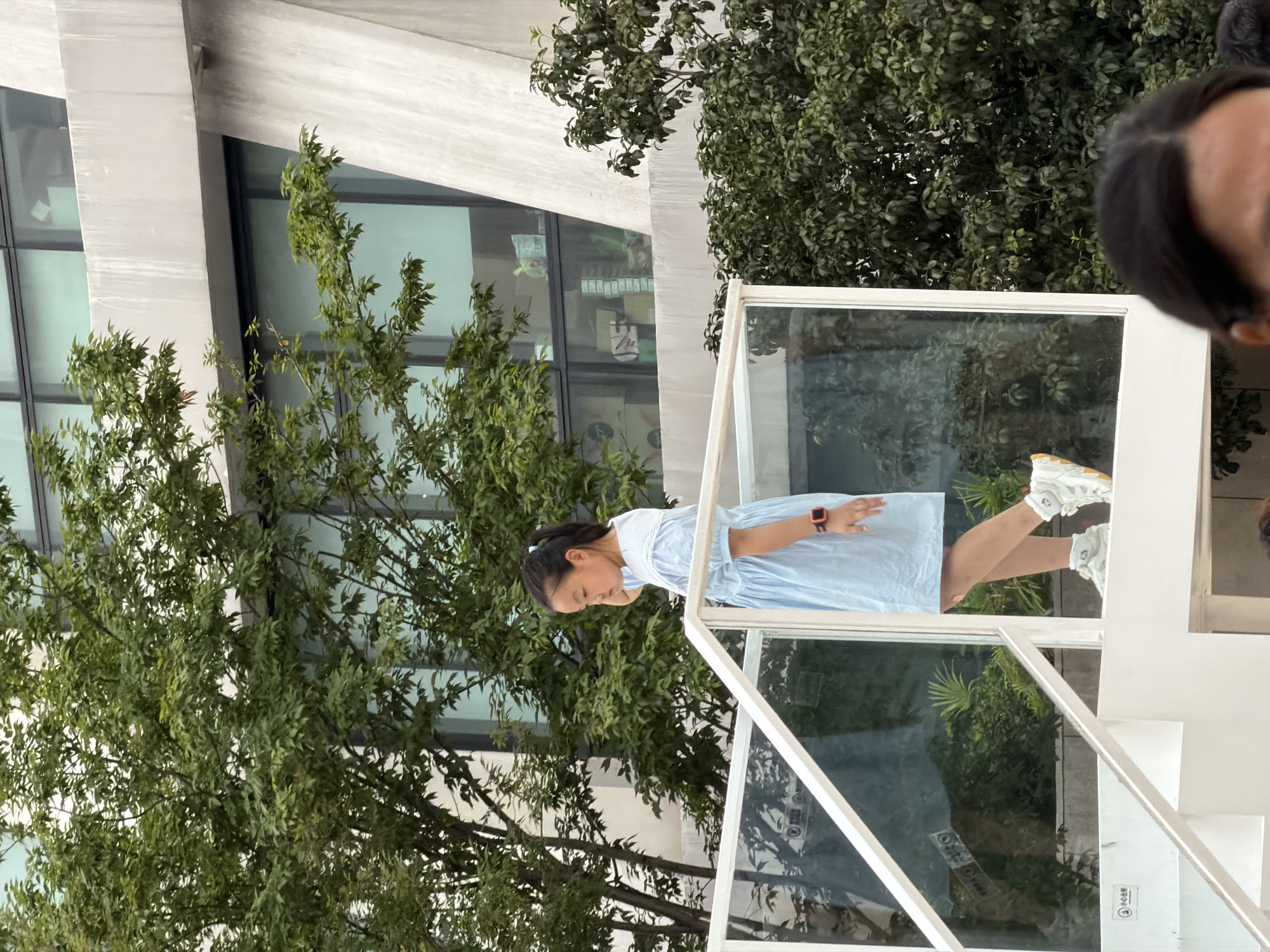 In Yuzhong District, China, a young East Asian girl with dark hair pulled back in a ponytail, wearing a light blue dress and white sneakers with orange accents, is walking on a white, elevated walkway. She is looking down and to her left. A smartwatch with a red band is visible on her left wrist. The walkway has a glass railing on the left and a white railing on the right. Behind her, lush green trees partially obscure a modern building with large windows and geometric concrete architectural elements. The overall environment appears to be an outdoor urban setting with significant greenery.