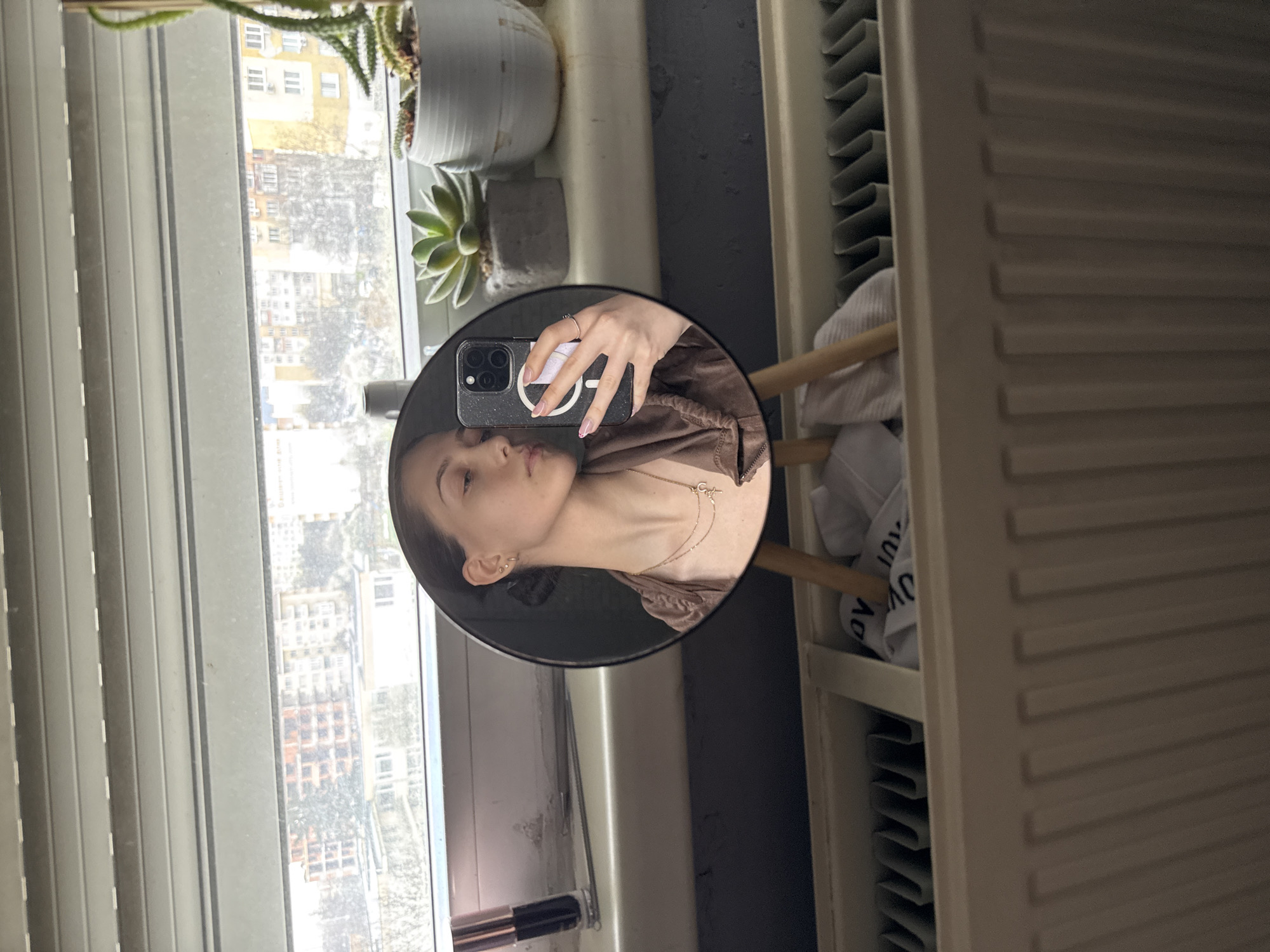 A woman is seen in a round mirror reflection, holding a phone with a pink and white phone grip on the back. She is wearing a brown hooded jacket and a necklace. The background behind the mirror contains a window with open blinds, revealing a cityscape with buildings. Green plants in pots are on the windowsill. Adjacent to the mirror is a white radiator. Given the architecture and urban landscape visible through the window, the location is likely Sofia, Bulgaria.