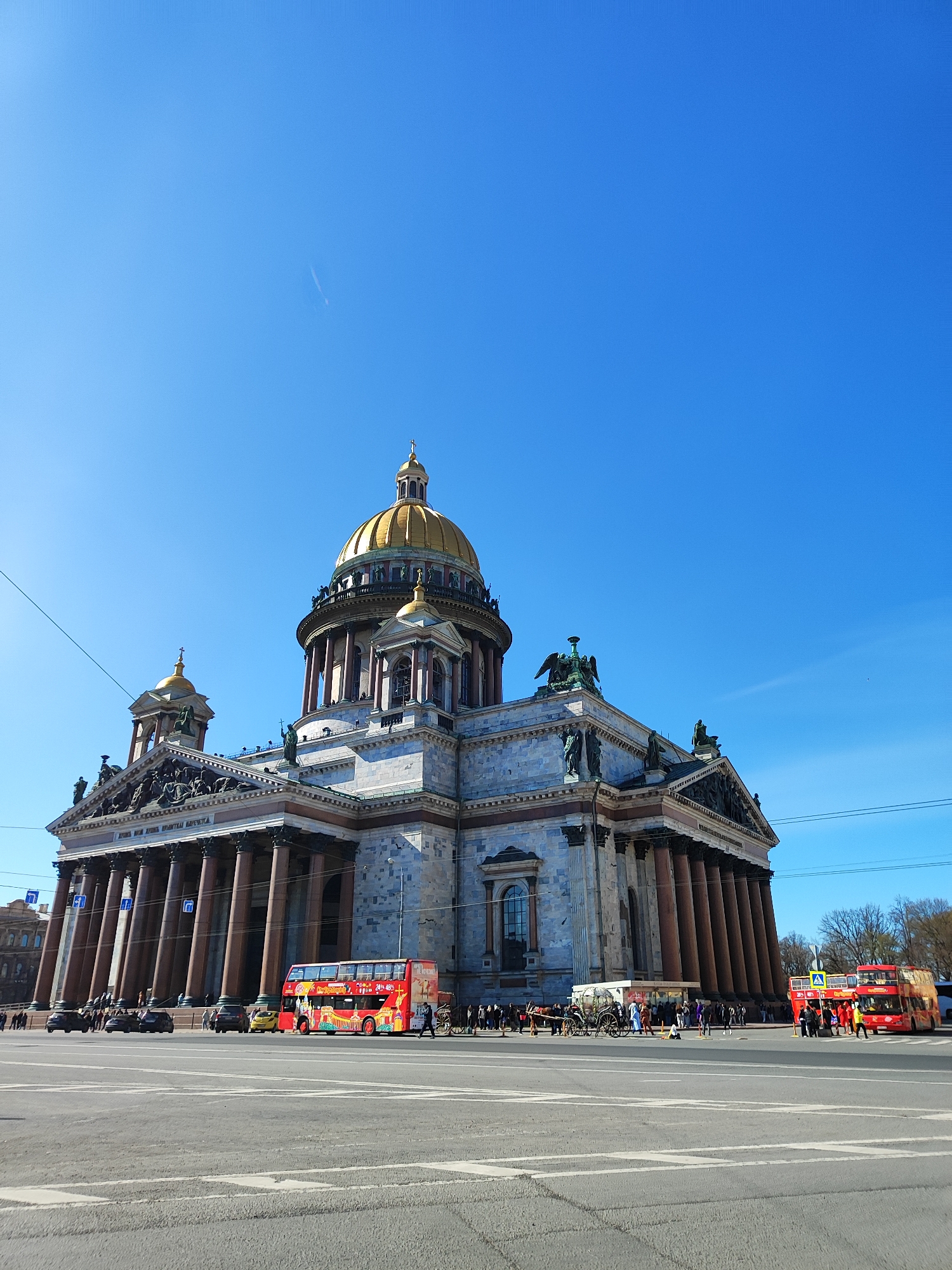 The image shows Saint Isaac's Cathedral in Saint Petersburg, Russia, under a clear blue sky. The cathedral, constructed of grey stone with a large golden dome, stands on a large square. A few electrical wires stretch across the sky in the upper left and right corners. In front of the cathedral, a variety of vehicles and people populate the square. Two red double-decker tour buses are visible, one on the left and one on the right. There are a few cars, and a horse-drawn carriage with people standing nearby. Pedestrians are walking on the square. The asphalt shows lane markings.