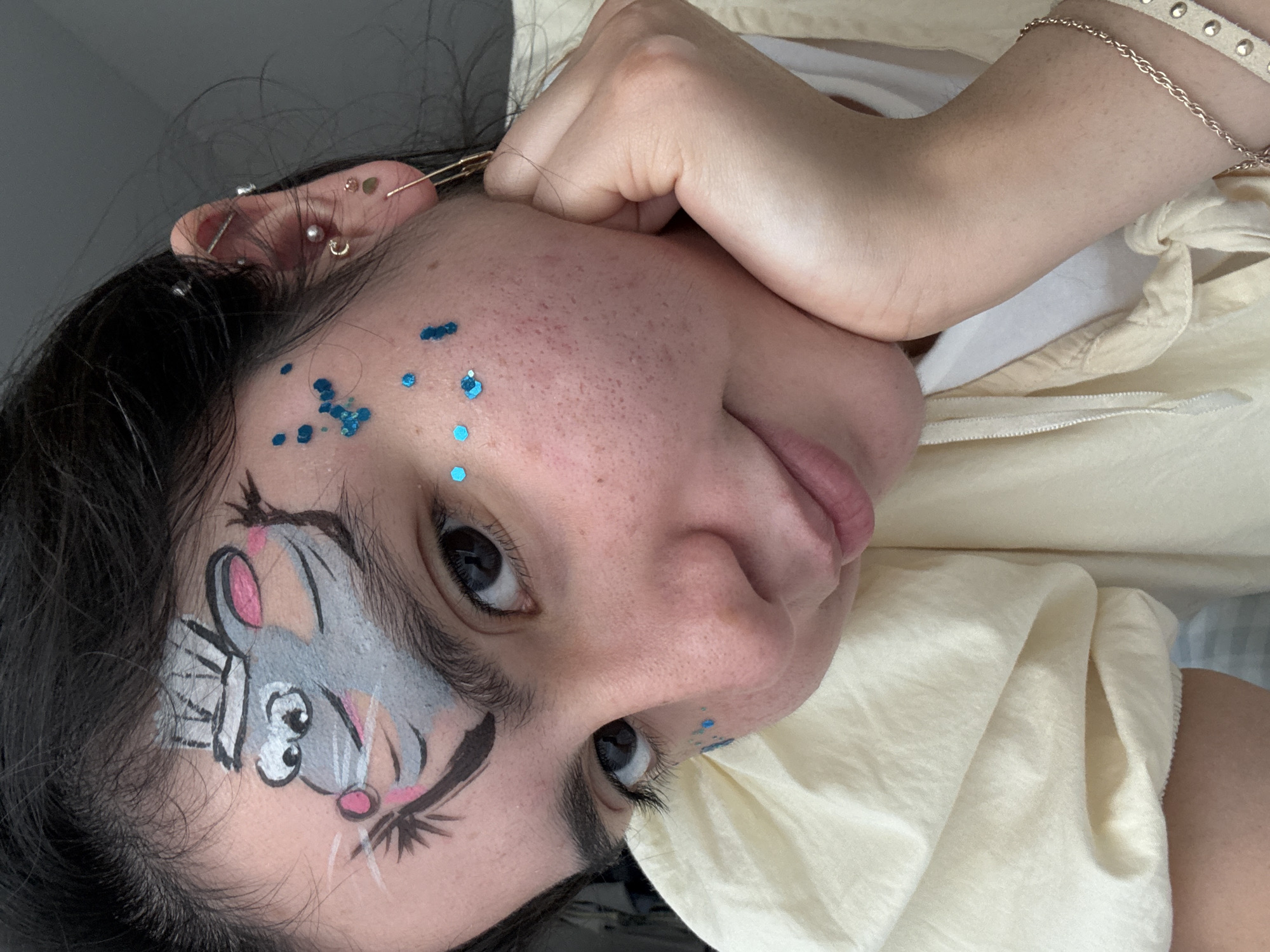 A young woman with dark hair is pictured from the chest up, resting her chin on her hand. She is wearing a light-colored, loose-fitting garment. Her forehead is adorned with face paint depicting a grey mouse wearing a chef's hat, with blue glitter scattered on her face. She has multiple piercings in her left ear. The background is nondescript, suggesting an indoor setting. There are no observable landmarks to place this image within the city of Austin, United States of America.