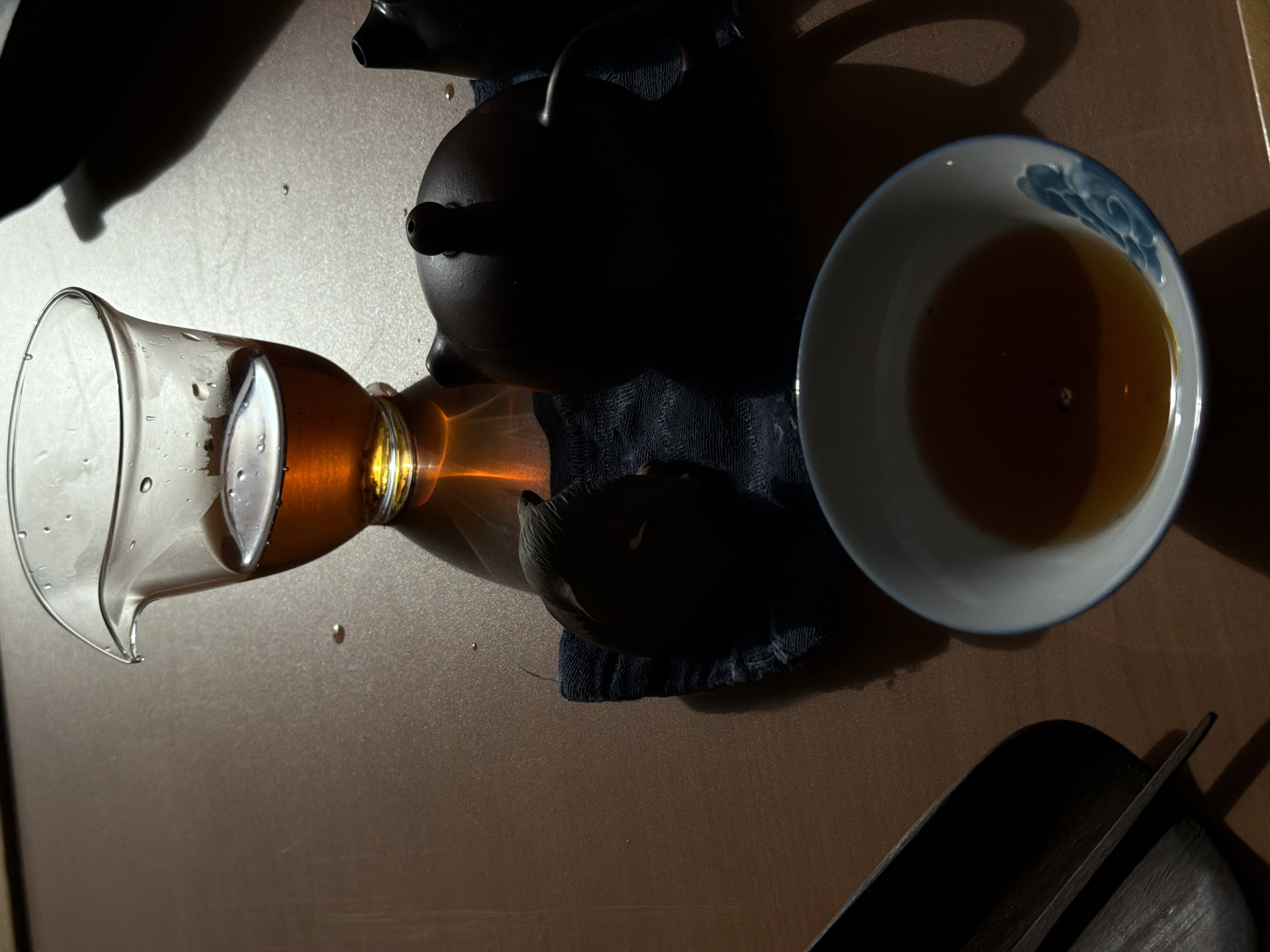 The image depicts a tea ceremony setup on a brown surface. A clear glass pitcher containing amber-colored tea lies horizontally on the left. Next to it is a dark-colored teapot resting on a dark blue cloth. To the right, a white and blue ceramic bowl filled with tea sits beside the teapot. A dark-colored object, possibly a tea tray or utensil, is visible in the lower right corner. Droplets of water are scattered around the pitcher. The scene is lit with strong light, creating sharp shadows. The image does not provide enough information to identify a city or location.