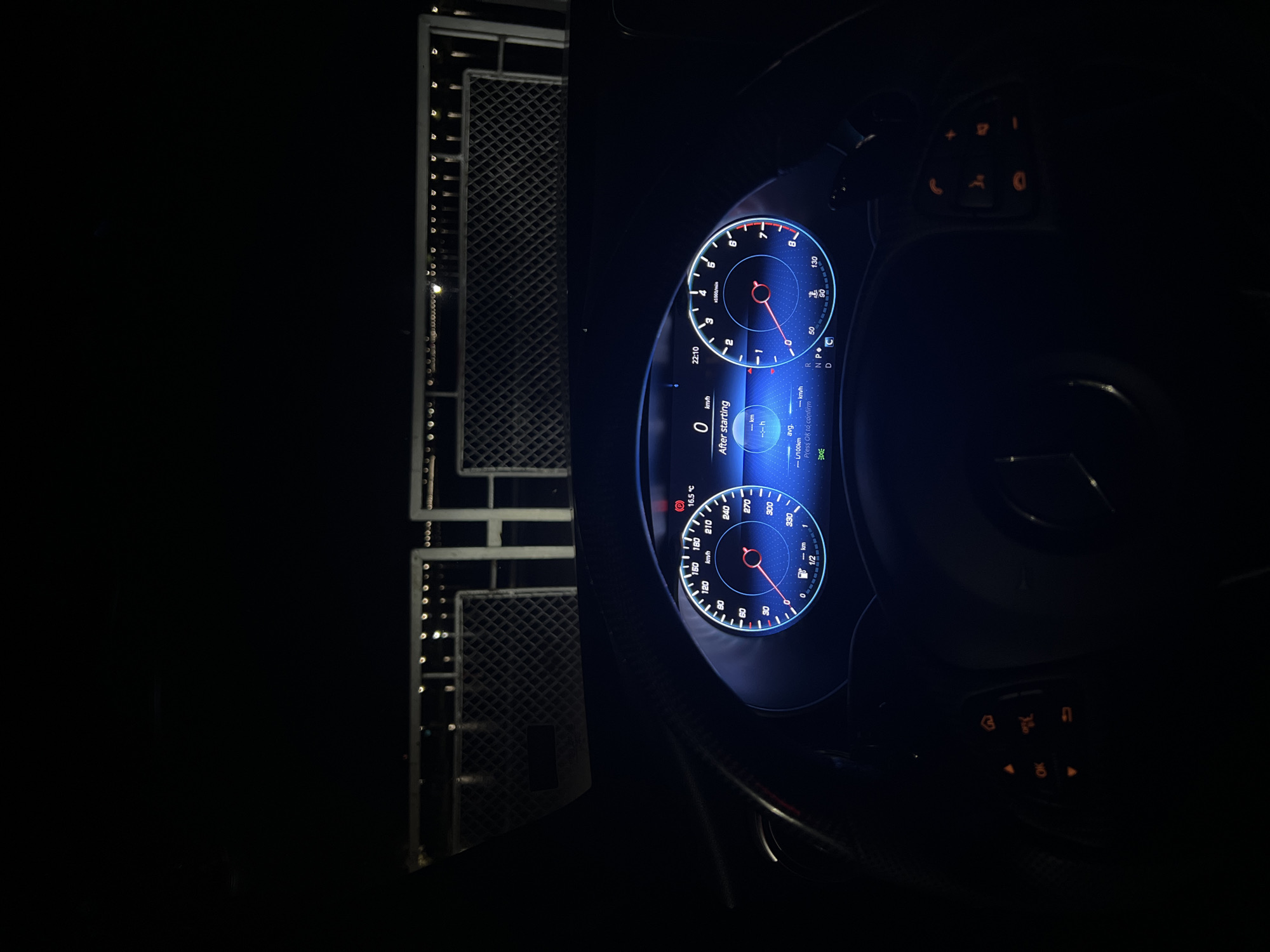 A nighttime scene inside a car, with the dashboard illuminated. The dashboard shows digital gauges displaying speed, RPM, fuel level, time (22:10), outside temperature (16.5 °C), and trip data "After starting" with 0 km/h. The speedometer shows a reading of 0 km/h and the RPM shows a reading of around 0. A steering wheel is visible in the lower part of the image. To the left of the dashboard, the image shows a blurred view of an exterior environment, possibly a metallic fence with embedded lights. The surrounding environment is dark. The image contains no identifiable information to determine that it is in the city of Plovdiv, Bulgaria.