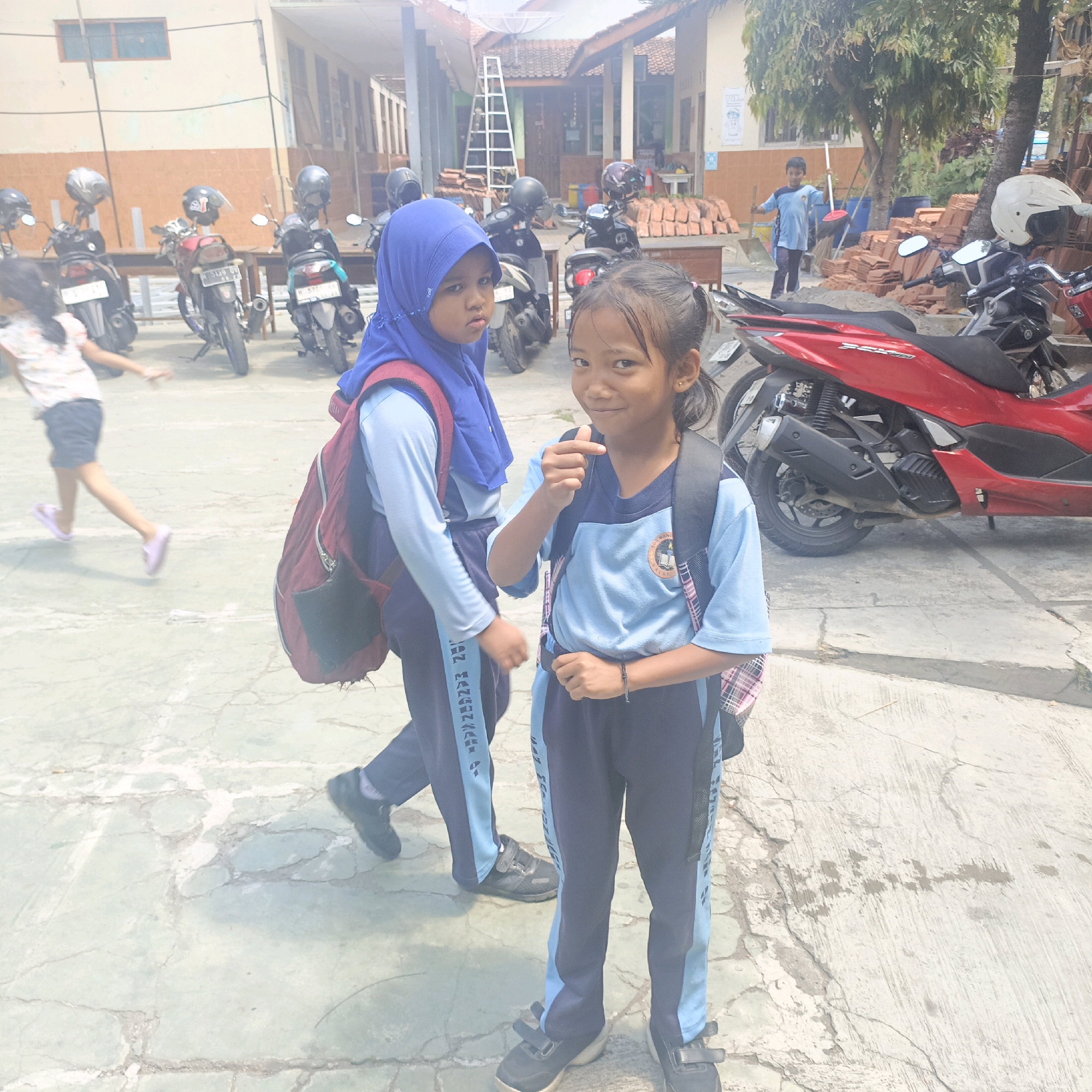 The image depicts an outdoor scene, likely a schoolyard in Salatiga, Indonesia, given the presence of uniformed children and parked motorcycles. Two young girls are the primary subjects. The girl in the foreground, facing the camera, has dark hair tied in a ponytail and is wearing a light blue t-shirt with a school emblem and dark blue pants with a light blue stripe. She is smiling and making a gesture with her right hand. She is also wearing a dark backpack. To her left, another girl, partially visible, is wearing a blue hijab and a similar uniform with a maroon backpack. In the background, several motorcycles are parked, some with helmets on them. A ladder leans against a building, and piles of bricks are visible, suggesting possible construction or maintenance work. The ground is paved with concrete. A young boy in a blue shirt is also visible in the mid-ground holding a broom and a shovel. The overall lighting suggests a bright, sunny day.