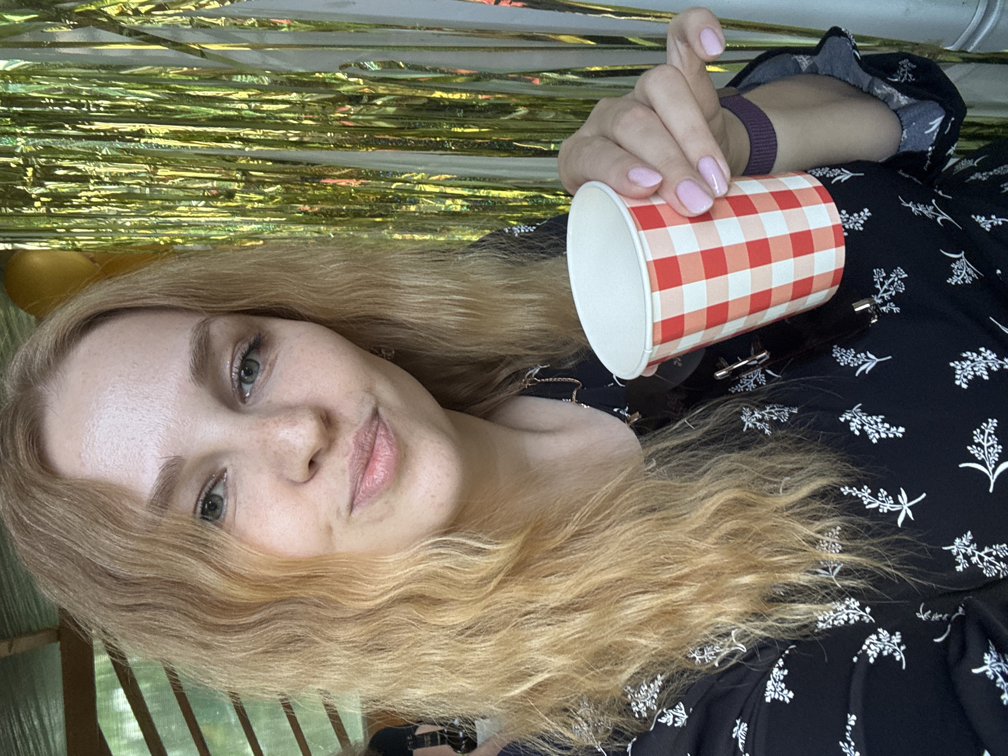A fair-skinned adult female with long, wavy, blonde hair and light-colored eyes is captured in a head-and-shoulders portrait shot in the city of Rozhdestveno, Russia. She is looking directly at the camera with a subtle smile, her lips slightly parted. Freckles are visible across her nose and cheeks. She wears a black garment adorned with a white floral or botanical pattern, and a faint metallic necklace is visible. Her left hand, with light pink painted fingernails, holds a red and white checkered paper cup towards the viewer. A dark purple elastic band is on her right wrist. The background on the left features a partially visible wooden and translucent roof structure, with green foliage suggesting an outdoor setting. The background on the right consists of a golden metallic tinsel curtain, with a yellow balloon partially visible behind it, indicating a celebratory environment.