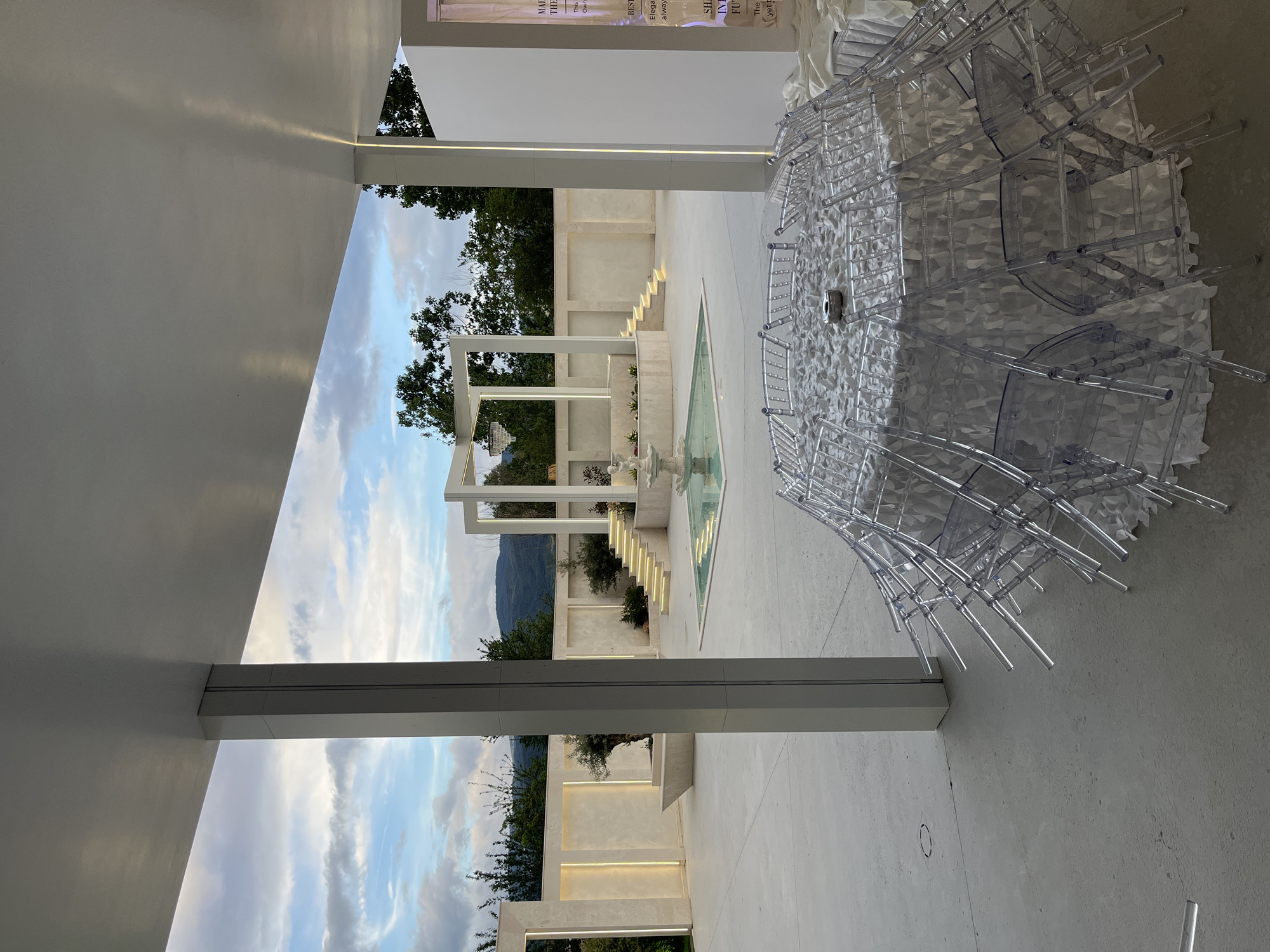 The image depicts an outdoor space with modern architectural elements. A stack of clear plastic chairs sits in the foreground on a concrete surface. In the middle ground, a geometrically designed structure with columns and beams frames a view of trees, a mountain, and a blue sky with clouds. A water feature, potentially a fountain, is visible within this structure. The architectural style is contemporary, with clean lines and a minimalist aesthetic. The location is Gotse Delchev, Bulgaria, based on the surrounding landscape.